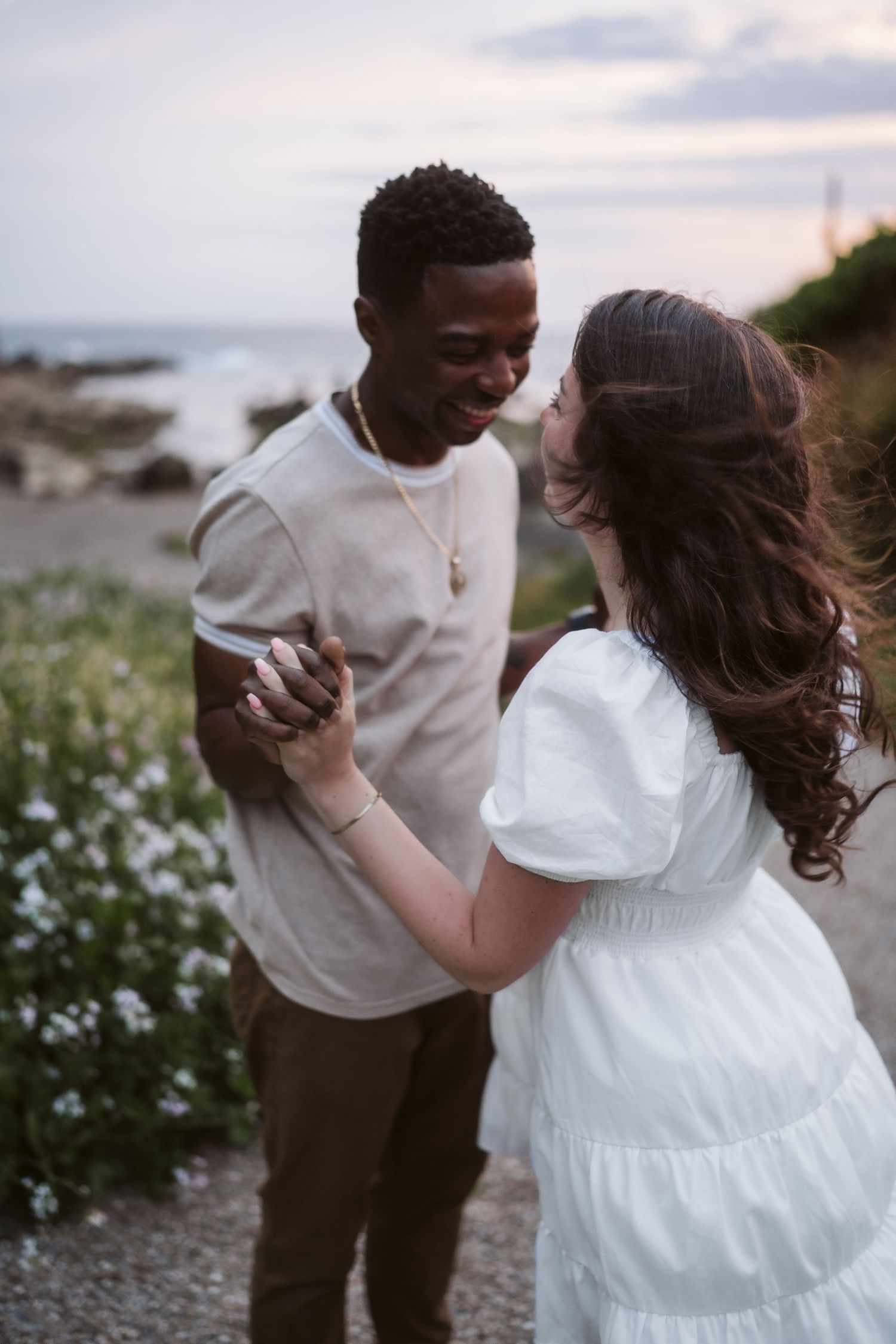 A couple dances together on a beach at sunset surrounded by coastal wildflowers.