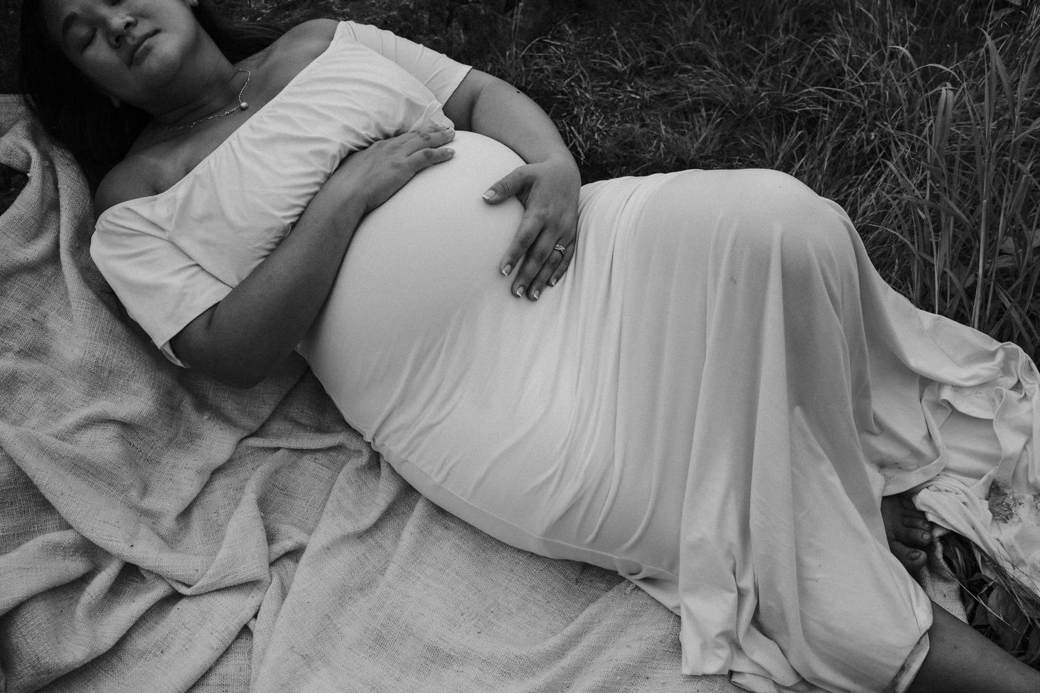 Black and white maternity photo of someone resting on a blanket in the grass wearing a flowing white dress.