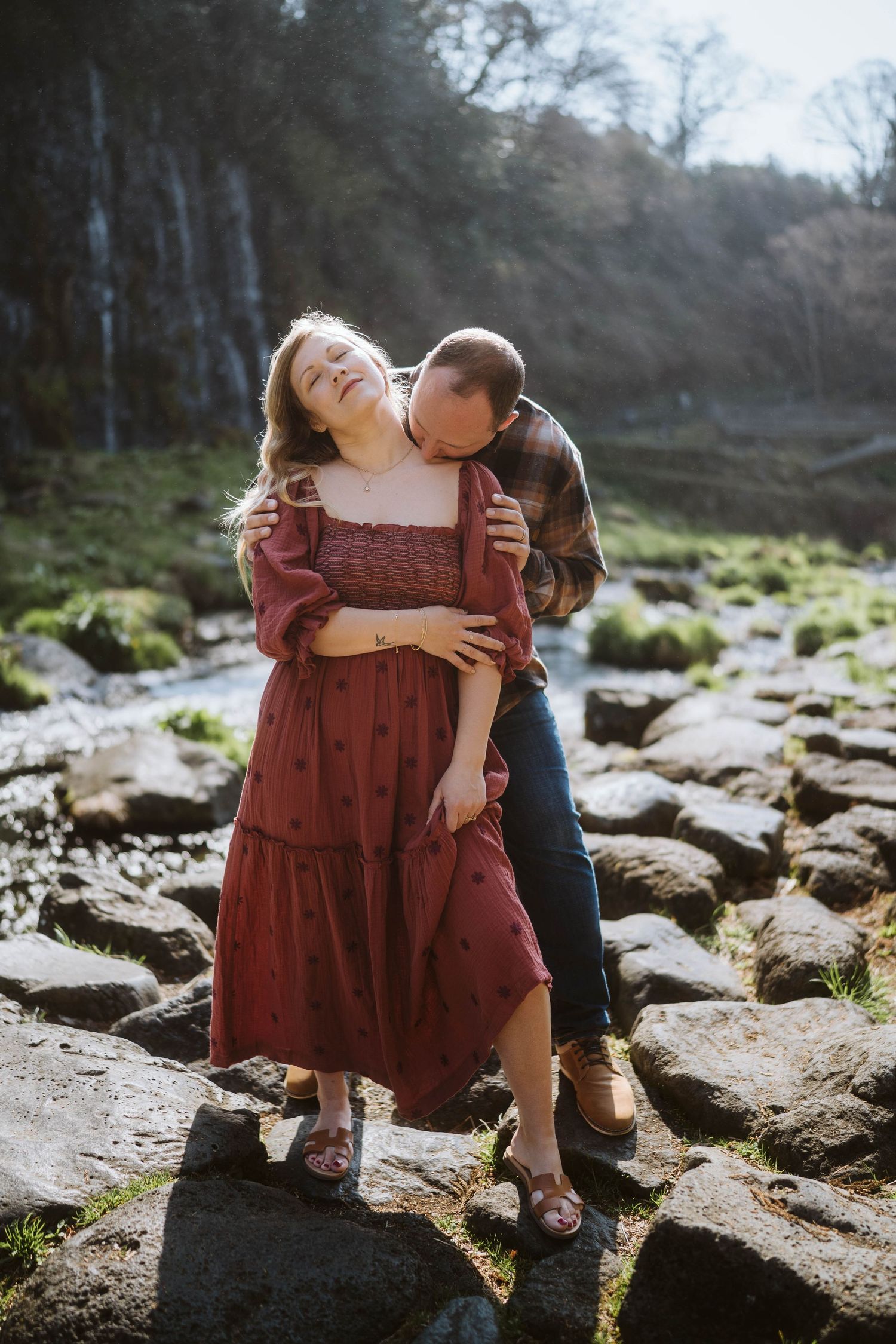 A couple shares a tender moment together while standing on mossy rocks near a waterfall on a sunny day.