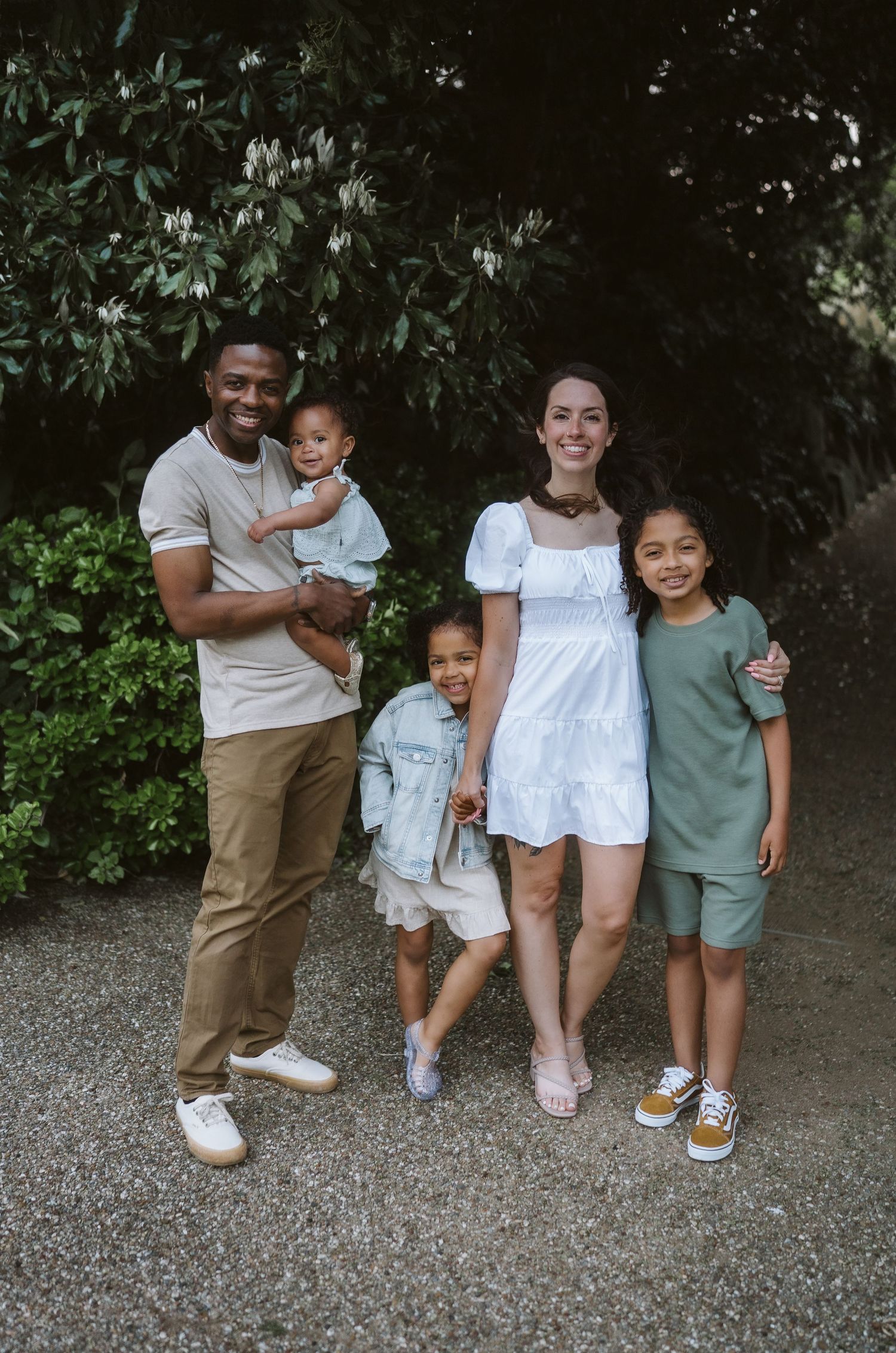 Family in casual attire stands together on gravel path with trees in background, showing candid loving moments.