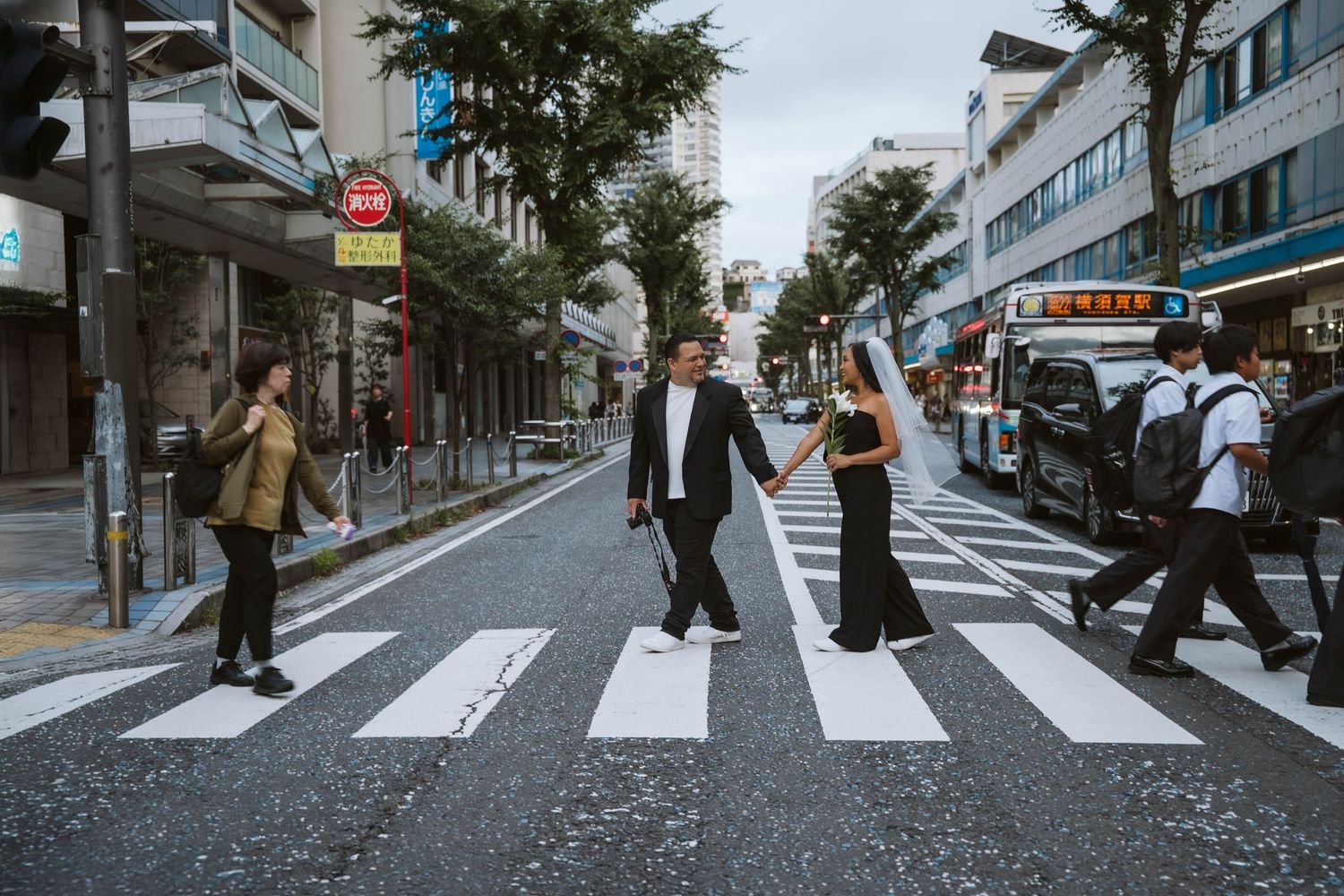 People walking across a crosswalk on a city street with buildings and trees visible in the background.