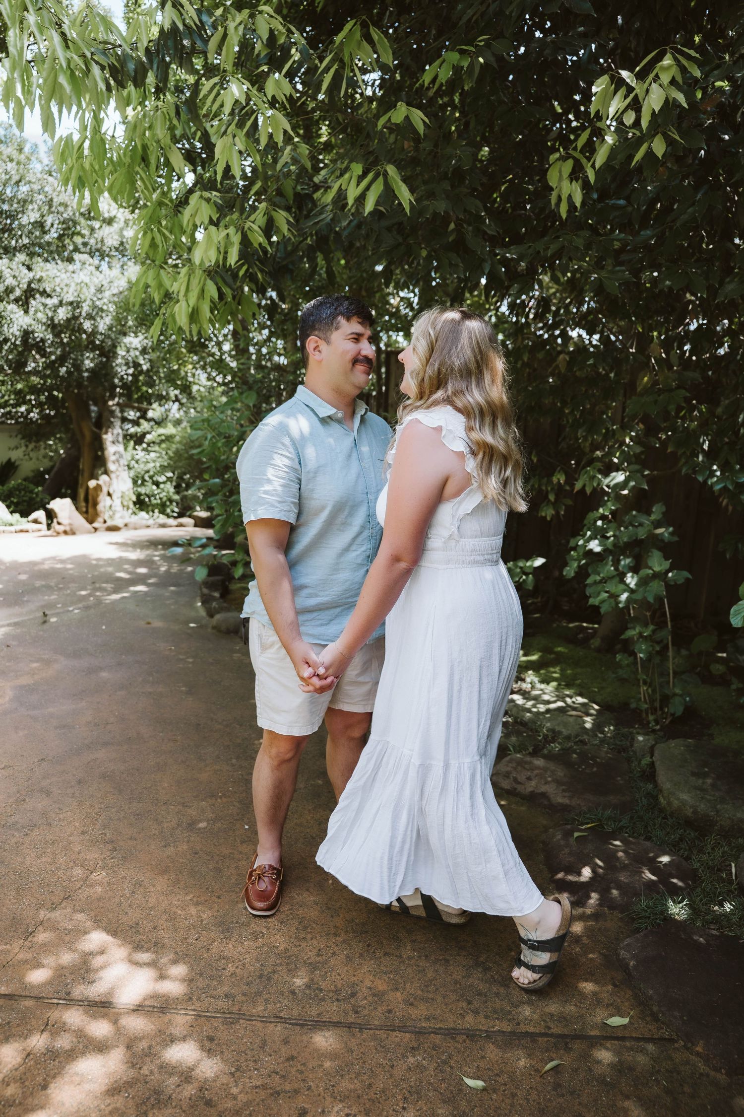 A couple shares a romantic moment on a garden path surrounded by lush greenery and dappled sunlight.