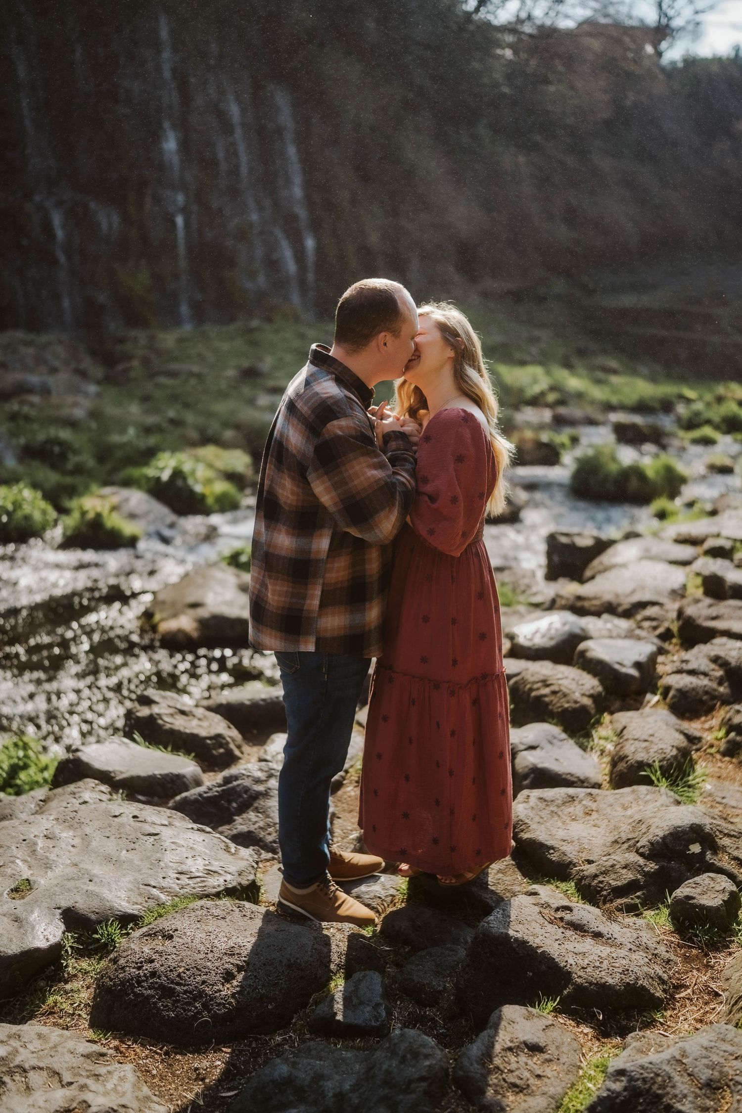 A couple stands together on rocky terrain during an outdoor photoshoot in a natural woodland setting.