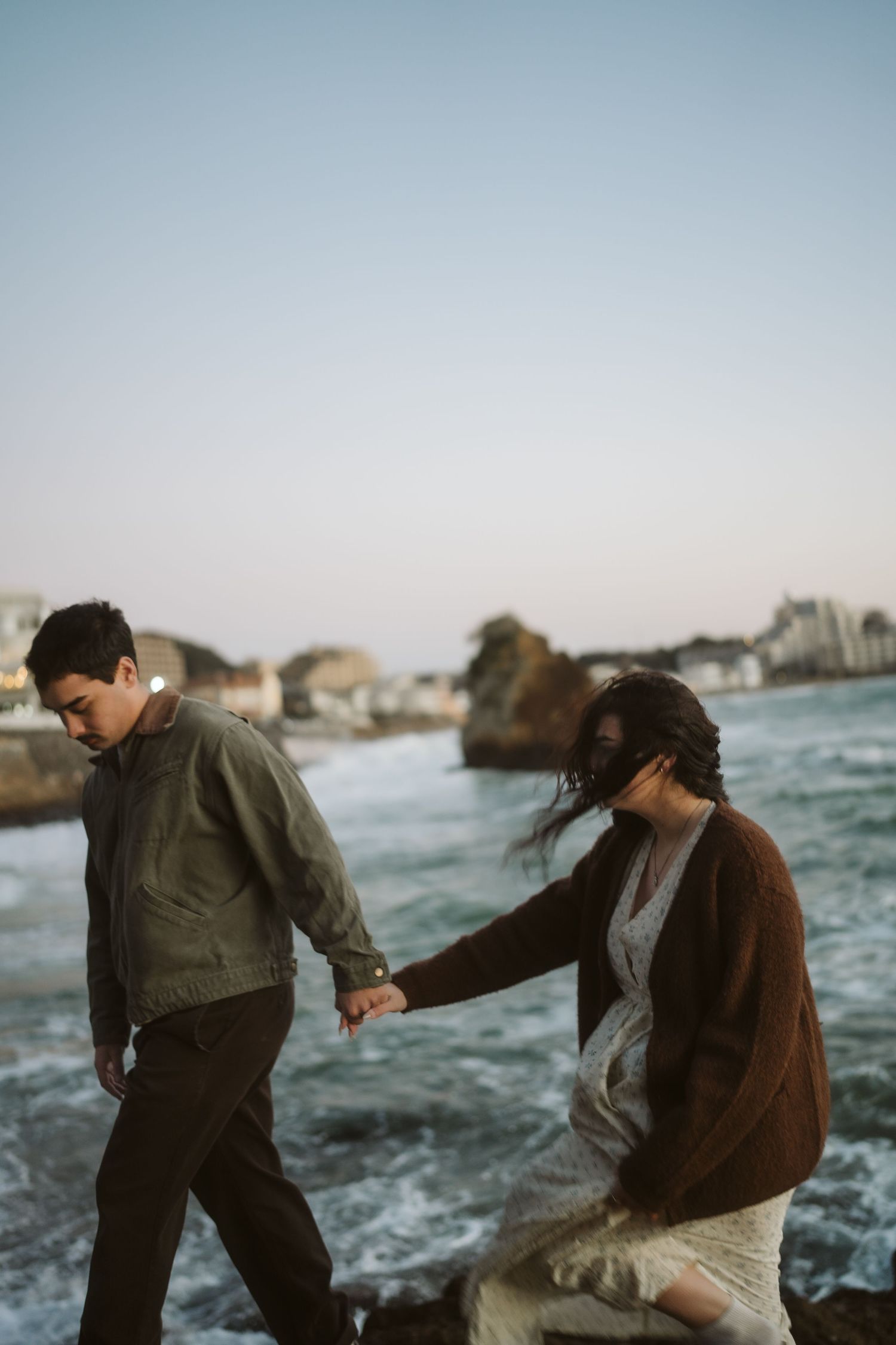 Two people dance and play together on a rocky beach at dusk with ocean waves behind them.