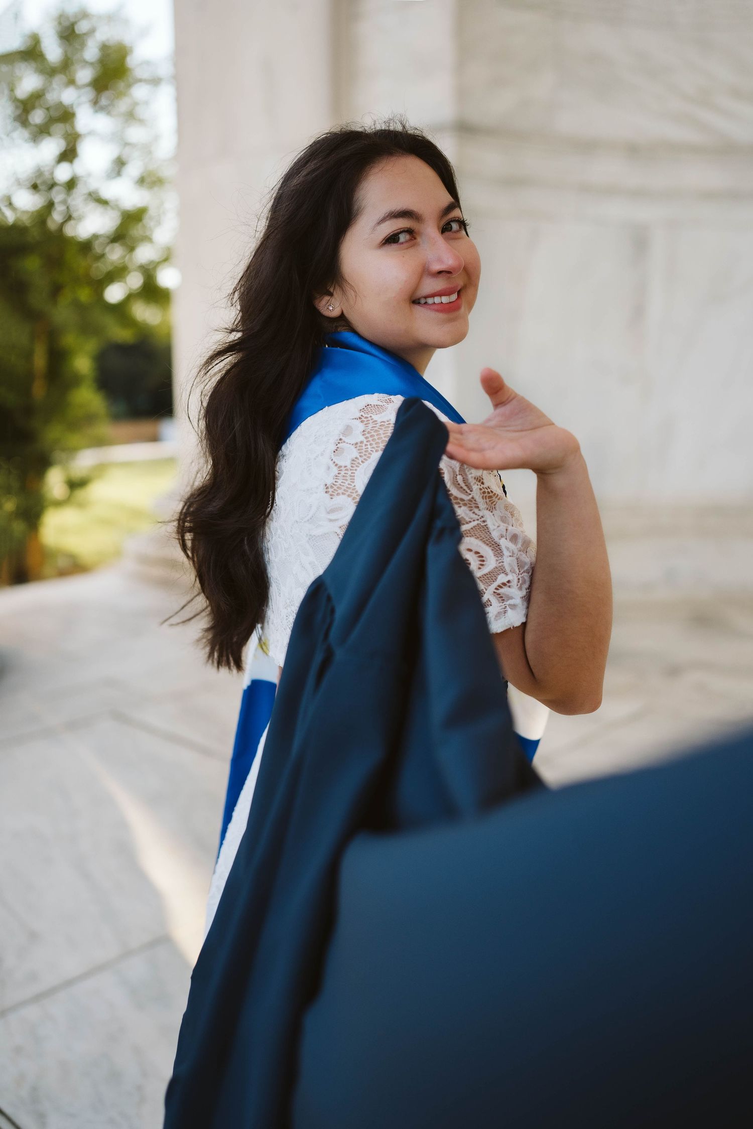 A graduate in a blue cap and gown turns to look back with a smile while wearing a white lace dress.