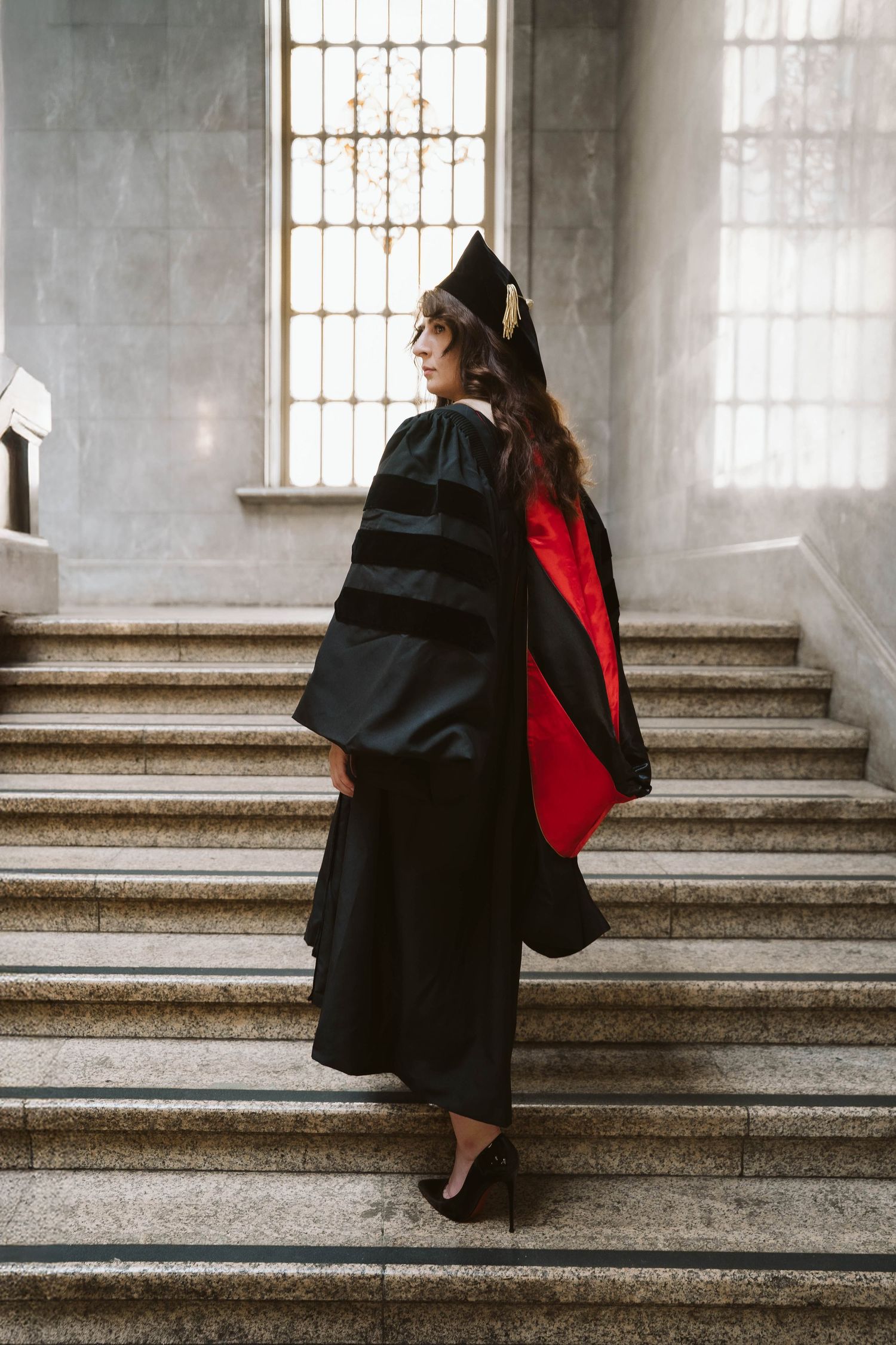 Graduate in black gown and red stole walking up grand stone steps in a historic building.