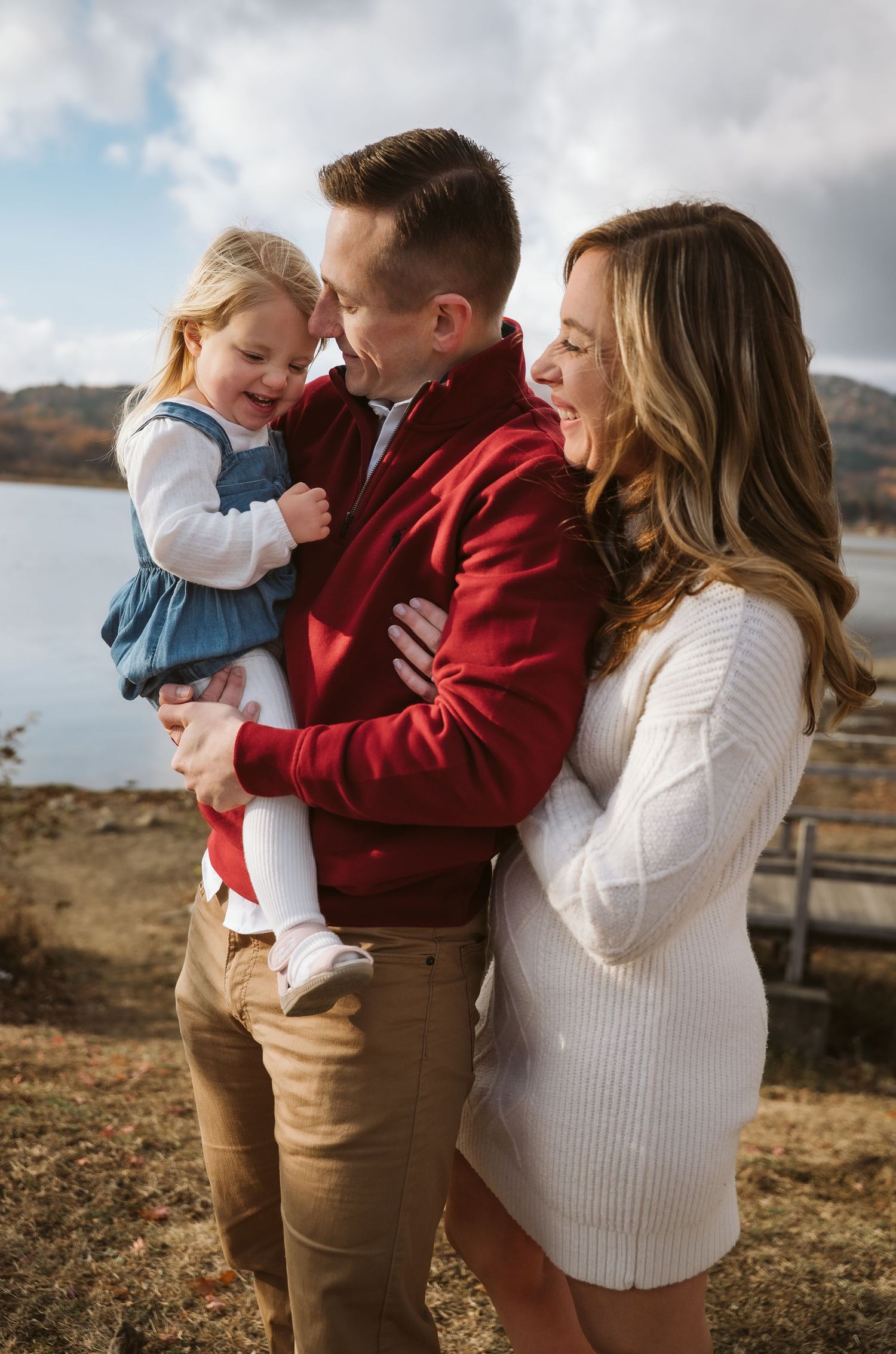Family wearing red and white sweaters embrace outdoors against scenic mountain backdrop during autumn photoshoot.