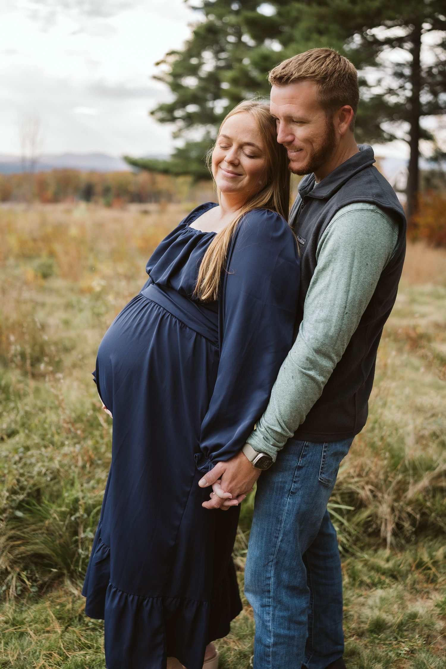 Couple standing in autumn field embrace as they await their new arrival in navy dress and casual wear.