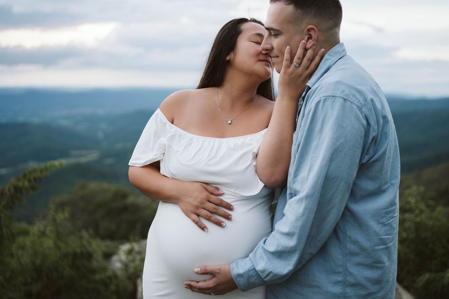 A couple embraces tenderly on a mountaintop overlook, with the woman wearing a white off-shoulder maternity dress.