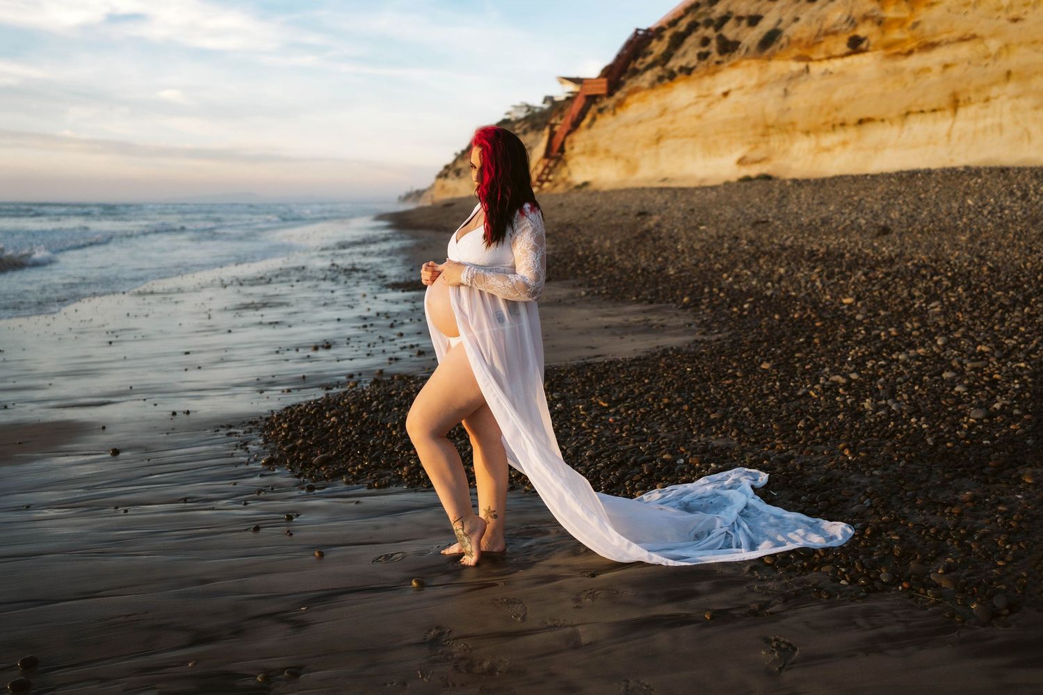 A flowing white dress contrasts against a rocky beach shoreline with dramatic cliffs at sunset.