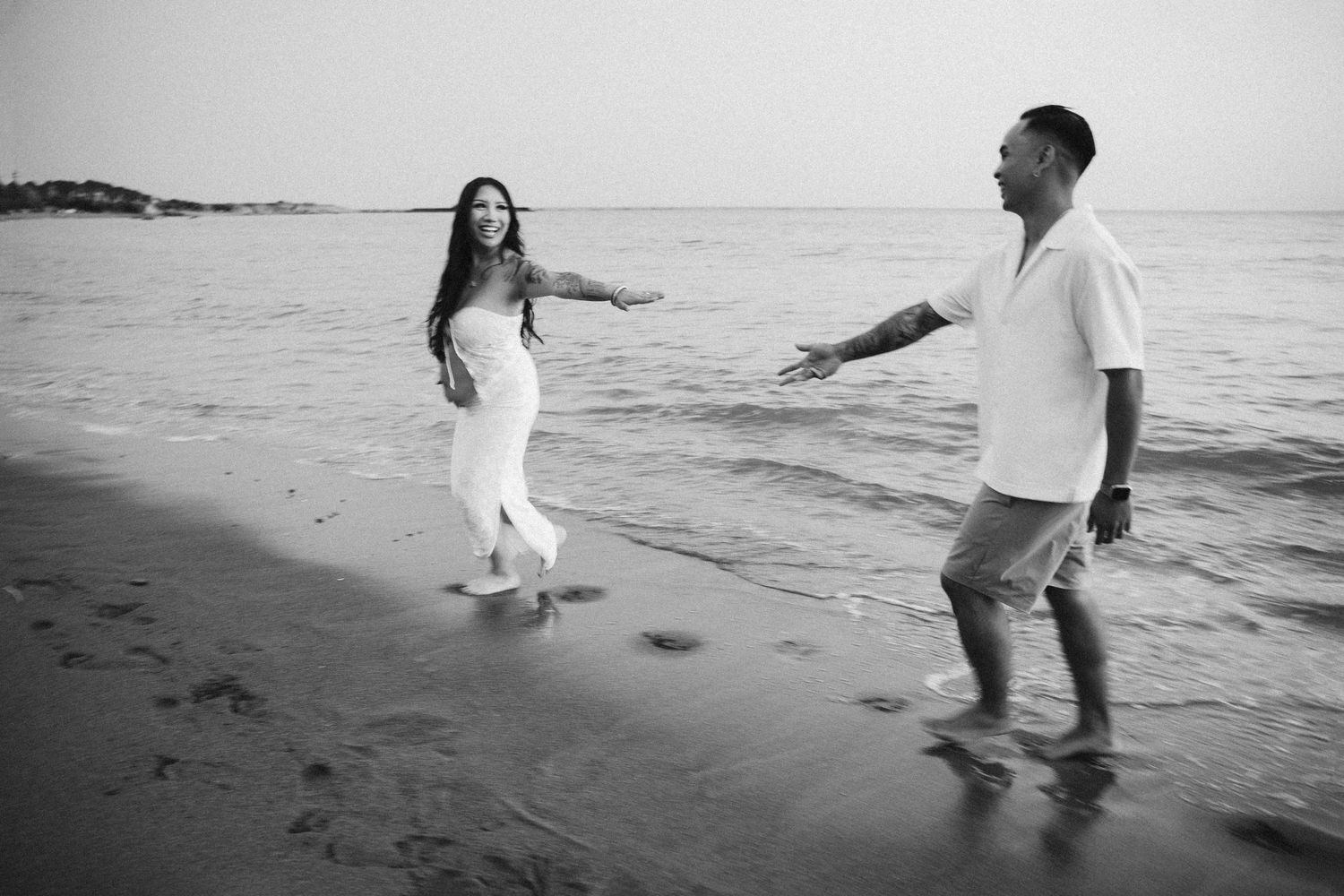 Black and white photo of a couple walking and playing along the shoreline at the beach.