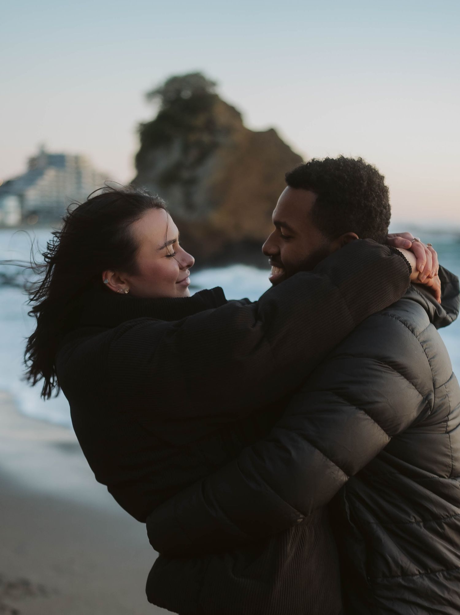 A couple embraces in silhouette against a moody sky during sunset at the beach.