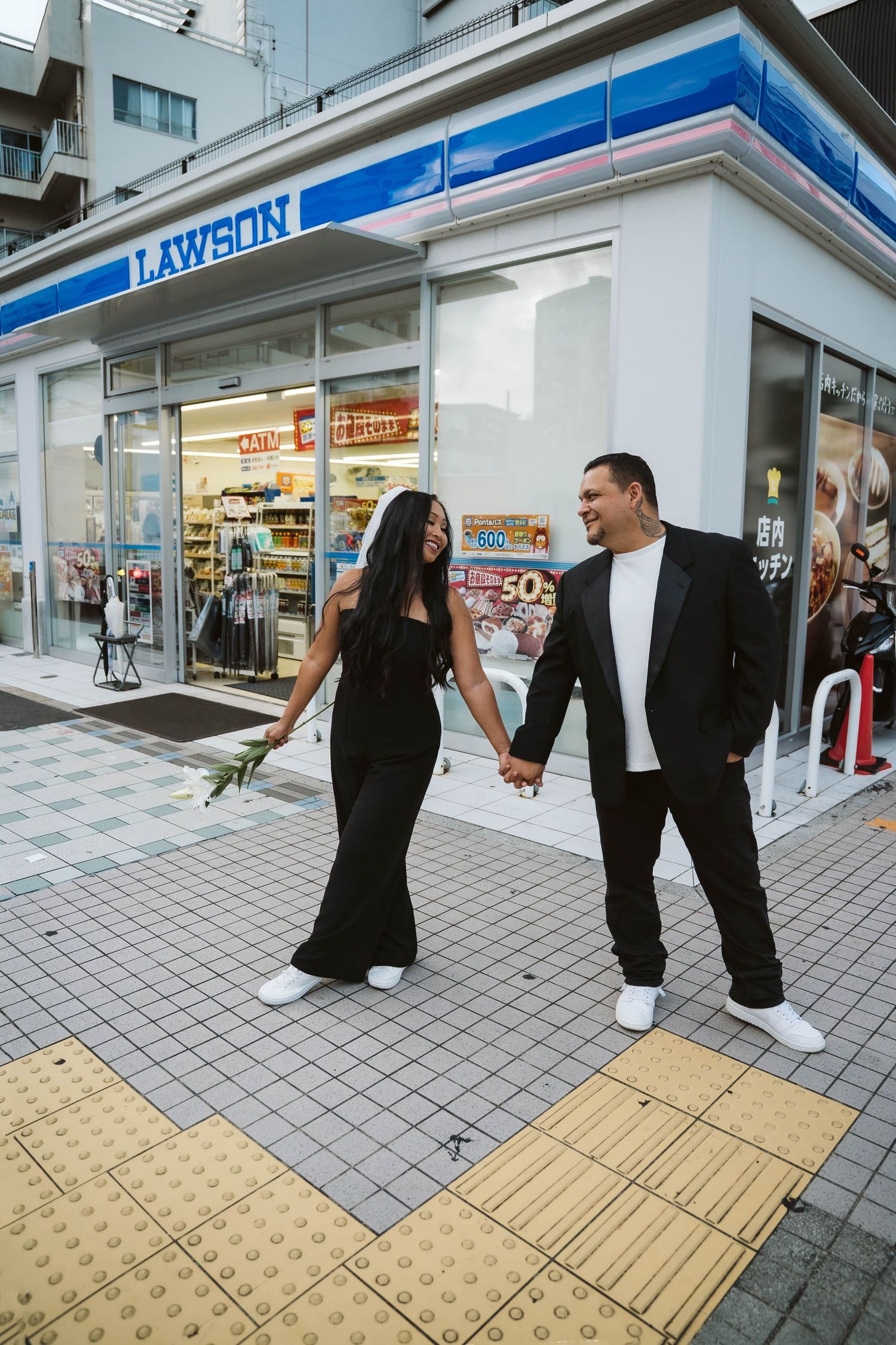 Two people in black and white outfits walking along a storefront sidewalk with yellow tactile paving.