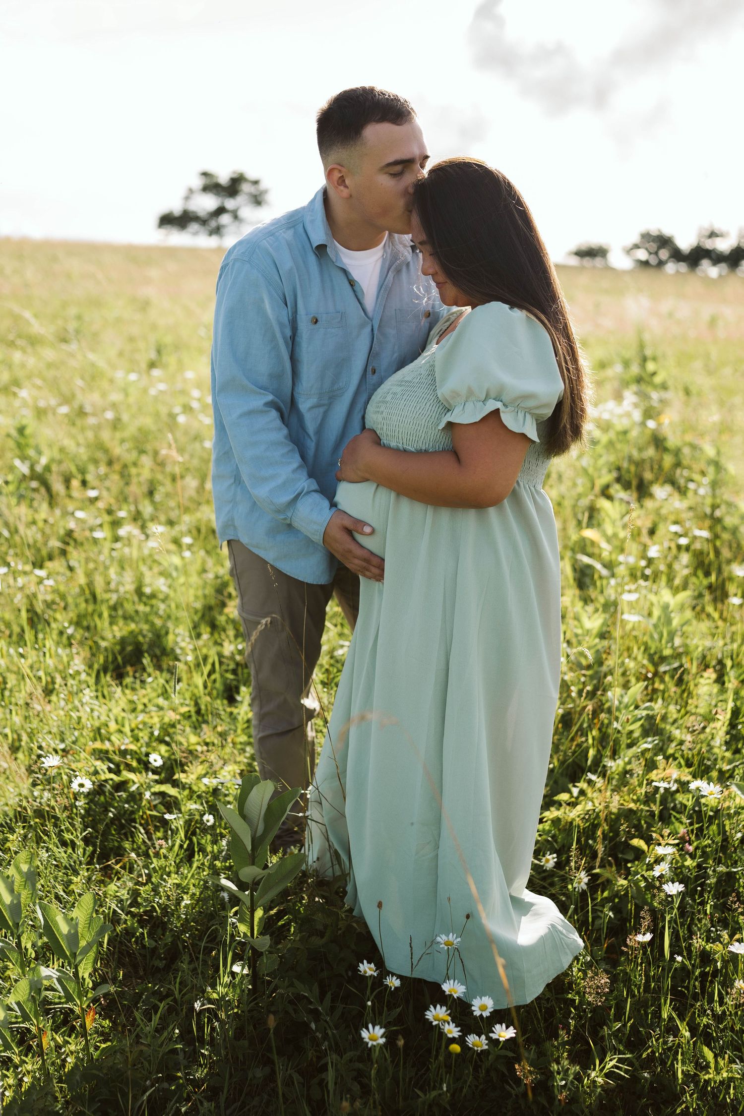 A romantic couple embraces in a sunlit meadow, with her wearing a flowing mint green dress and him in a blue shirt.
