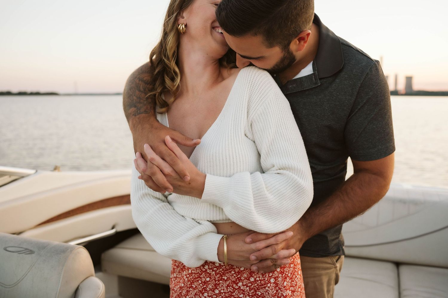 Romantic couple embraces on a boat during sunset, with person wearing white sweater and floral skirt.