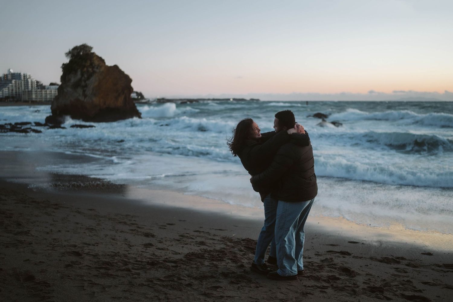 Couple shares intimate moment on beach at dusk with stormy ocean waves and rocky coastline.