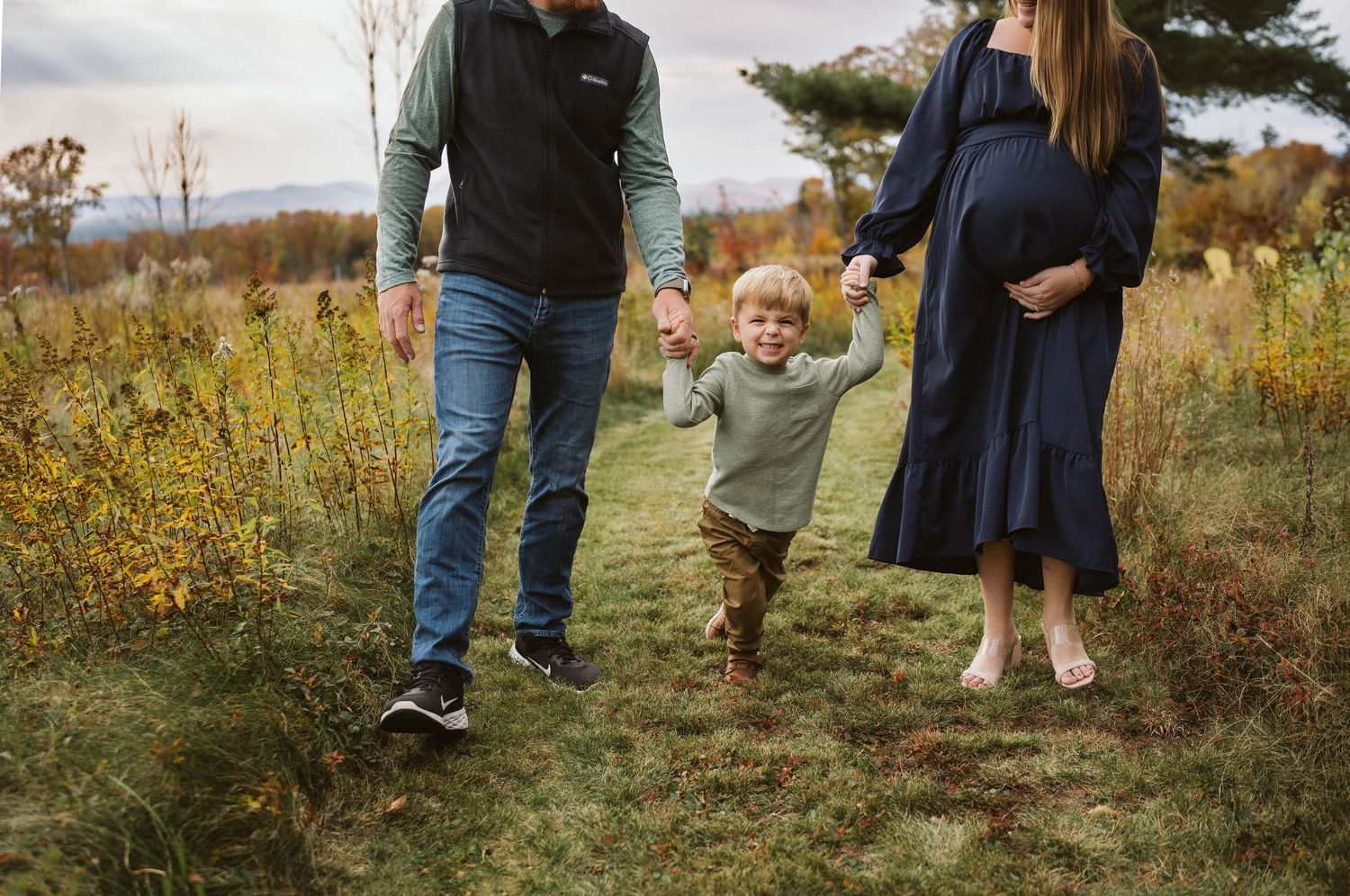 A family walks together on a grassy path through autumn foliage on a cloudy day.