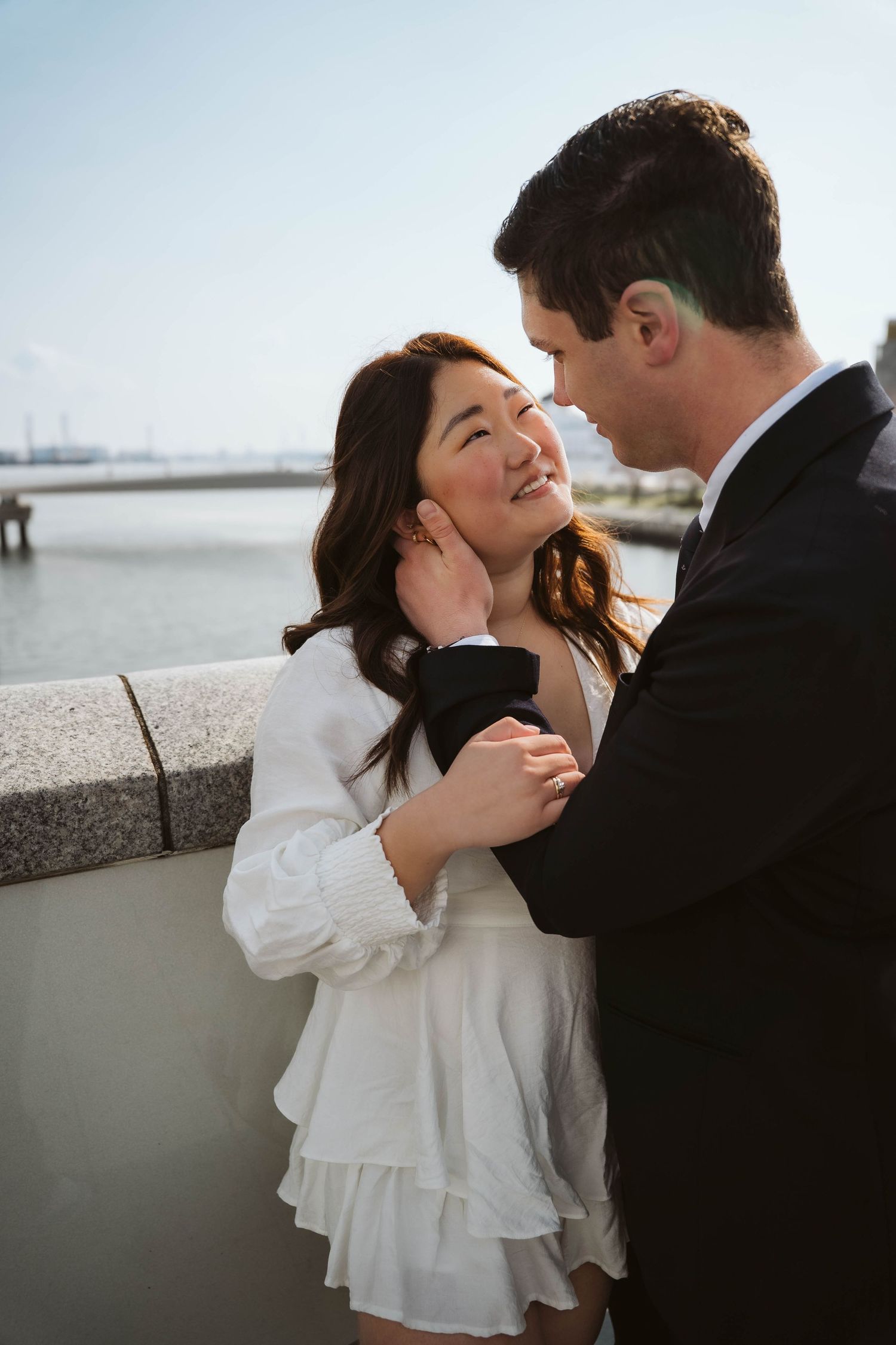A romantic couple shares an intimate moment by the waterfront, with her in a white dress and him in dark attire.