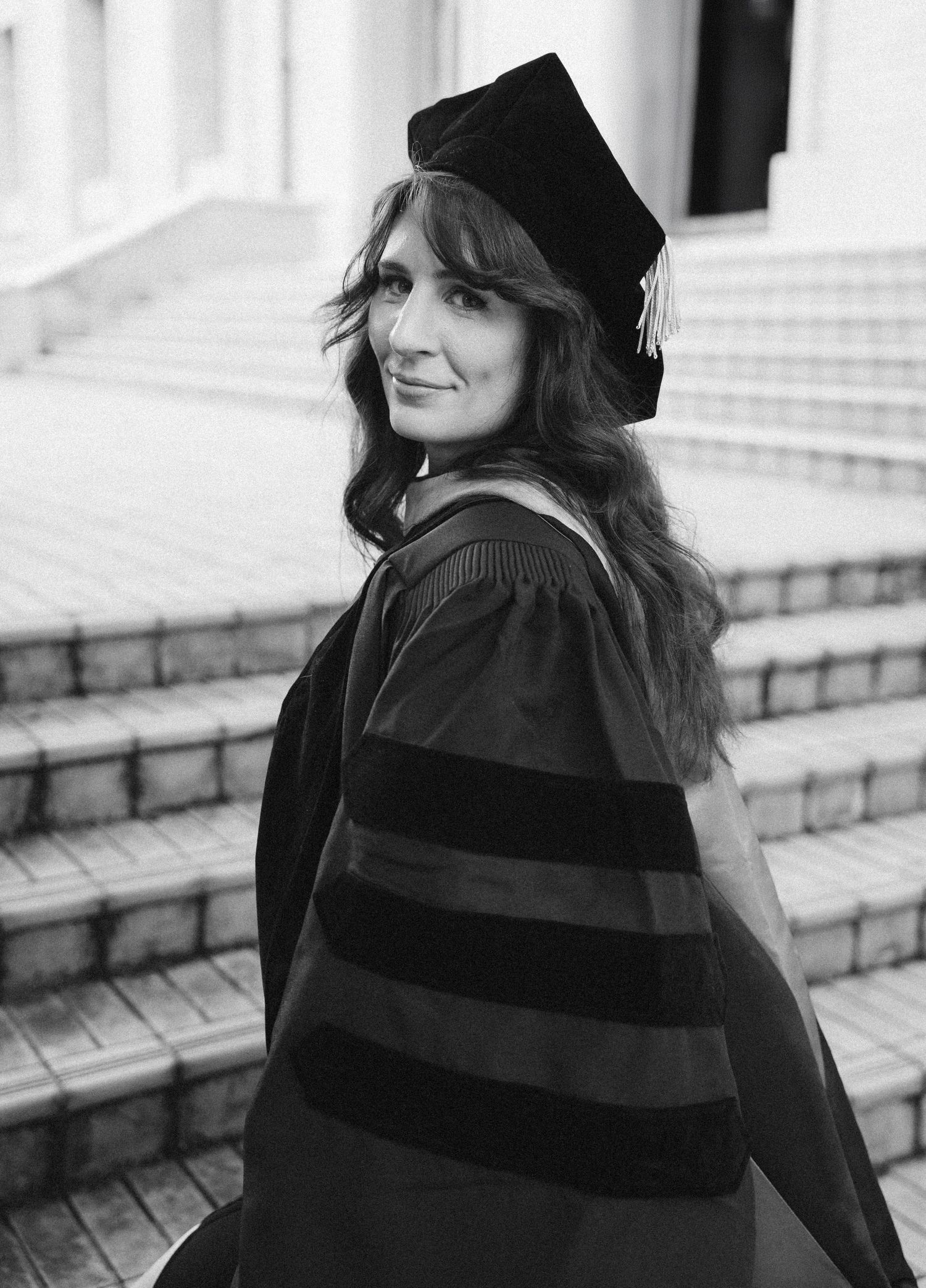 Graduate wearing traditional black academic regalia and cap poses on stone steps in black and white photo.
