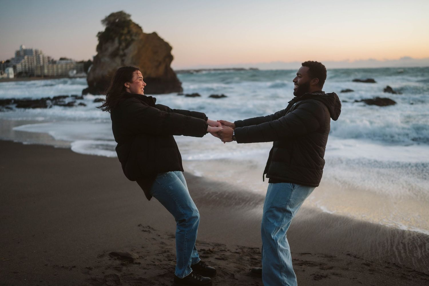 Silhouetted figures embrace on a scenic beach at sunset with crashing waves and rocky coastline in the background.