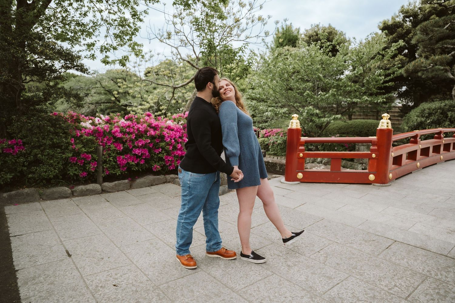 A couple stands on a red Japanese bridge surrounded by blooming azalea flowers in a peaceful garden setting.