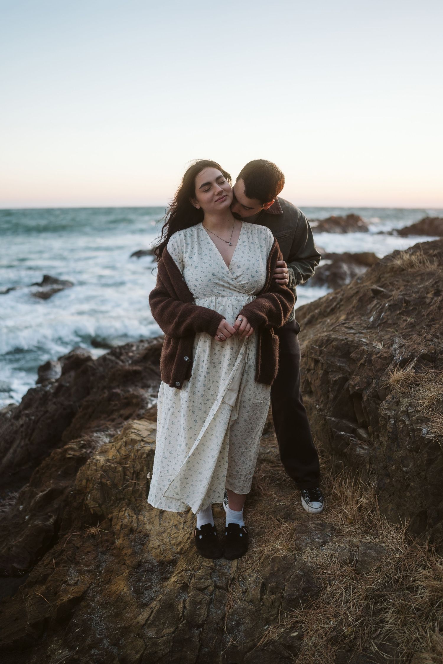 Couple sharing tender moments on rocky cliffs by the ocean during sunset.