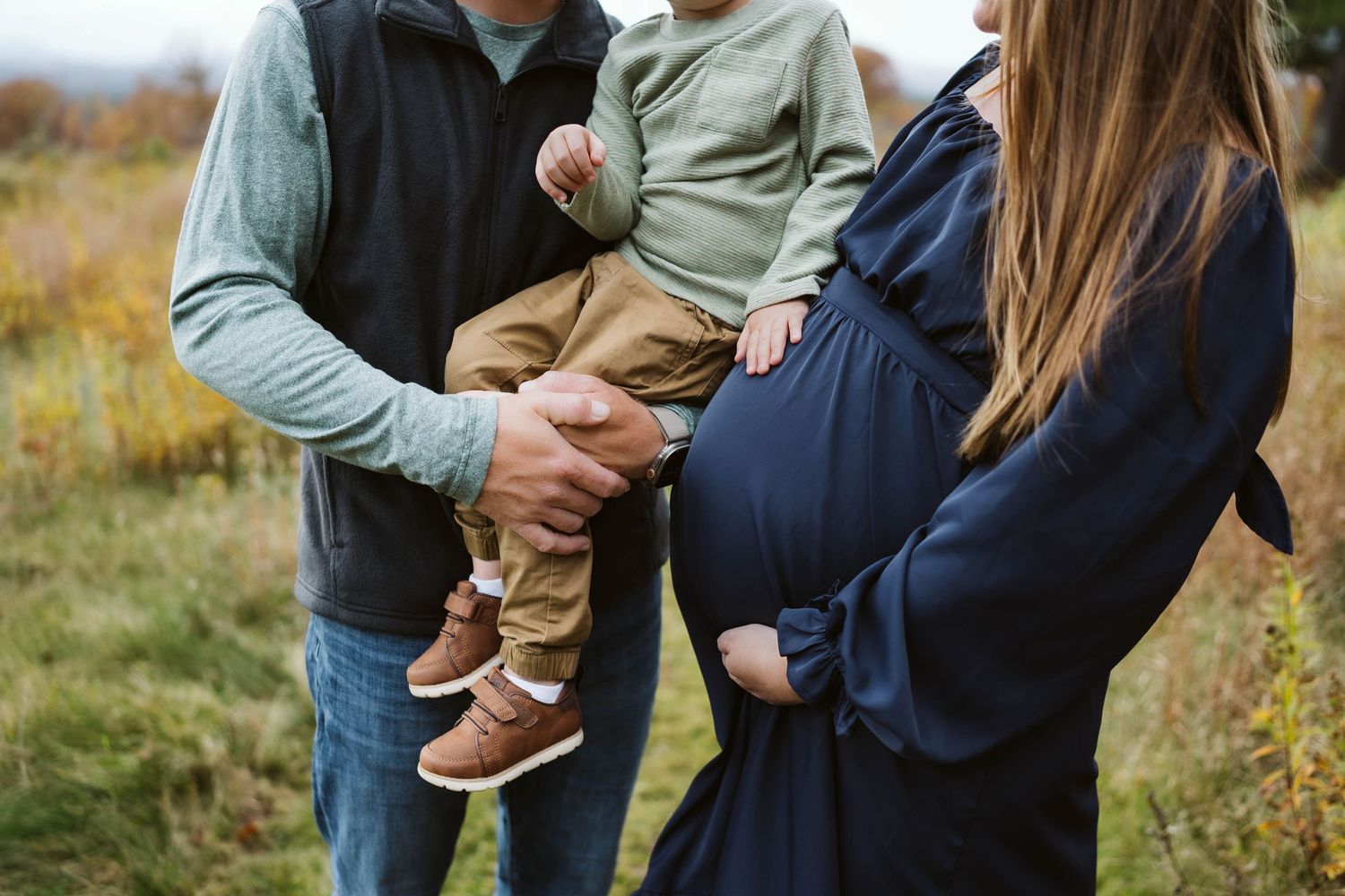 Expectant mother in navy dress with rounded belly stands next to family member holding toddler in outdoor fall setting.