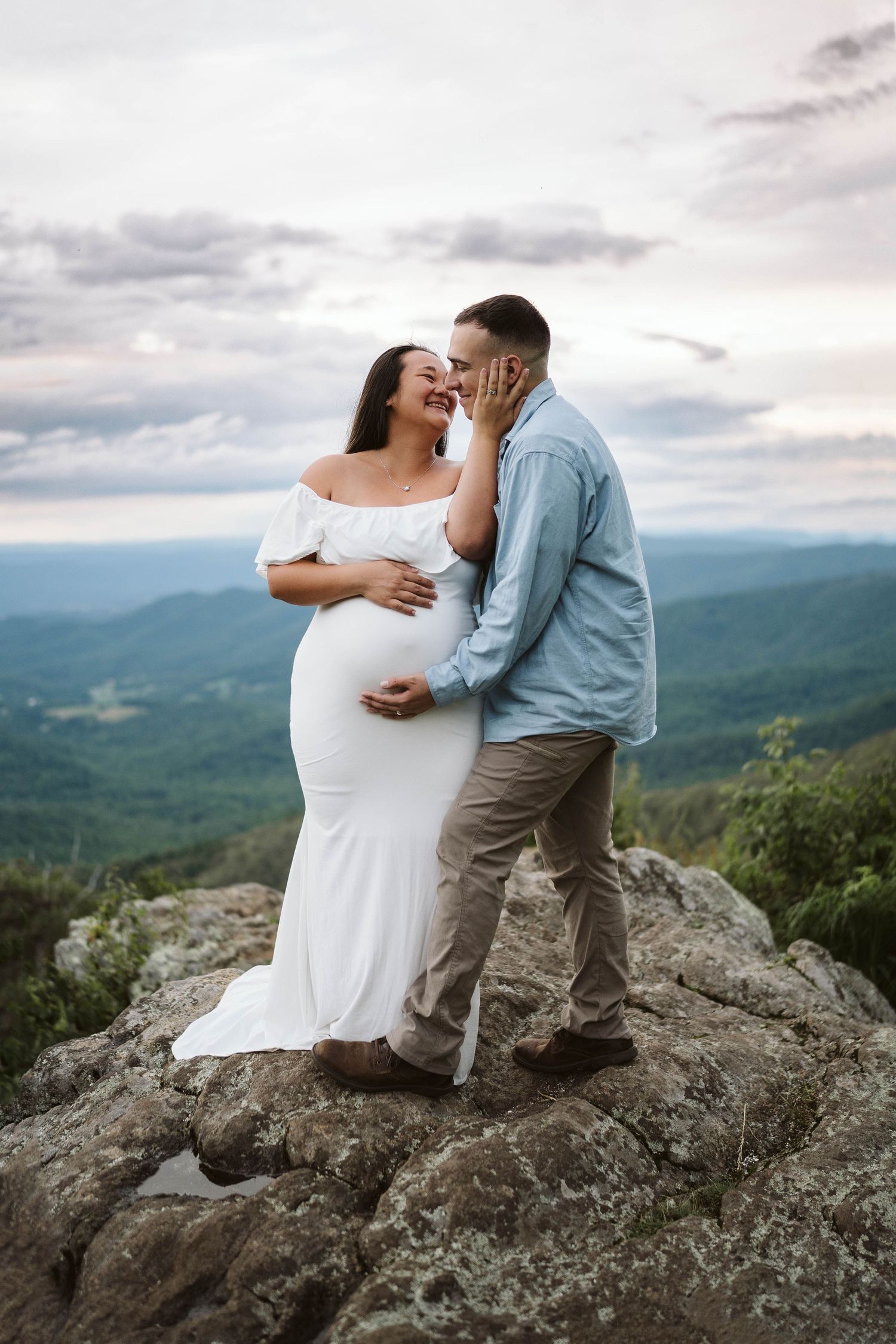 A couple shares an intimate moment atop a mountain cliff with scenic rolling hills and cloudy skies in the background.
