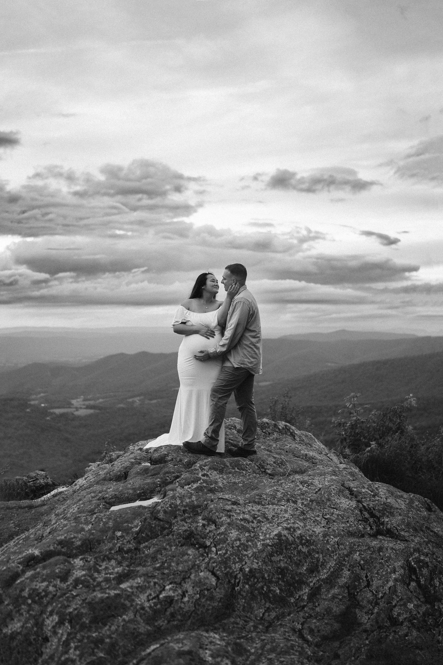 A couple embraces on a rocky mountaintop overlook with dramatic cloudy skies and rolling hills in the distance.