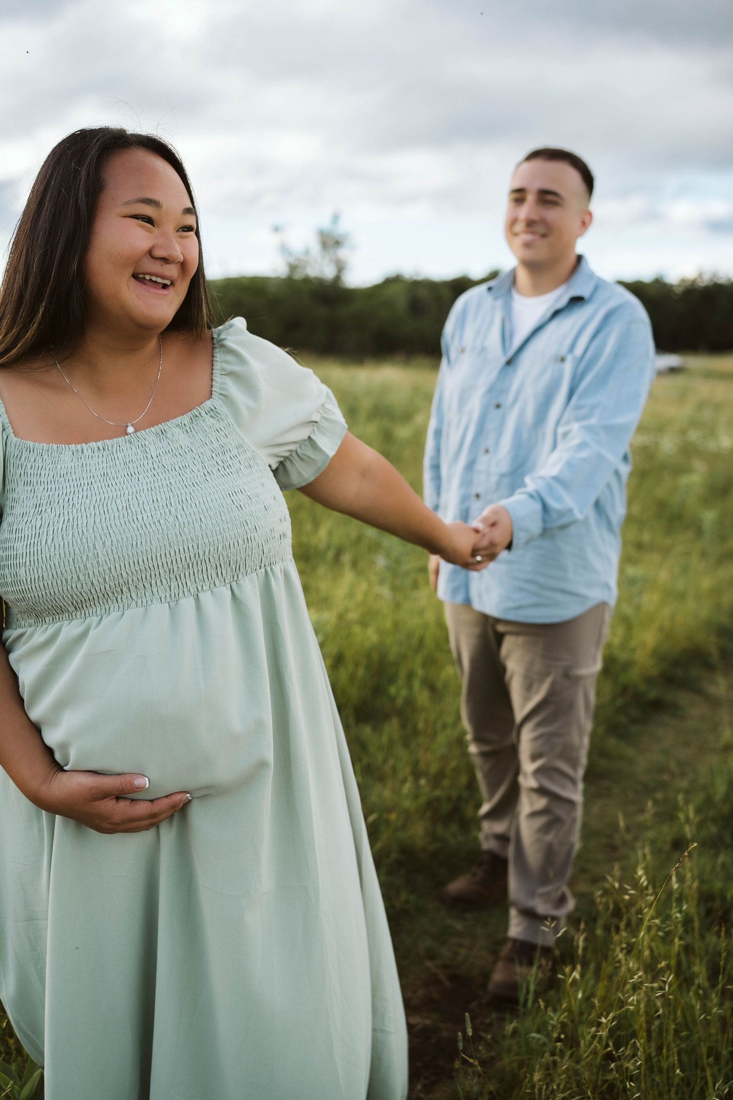 A couple stands together in a grassy field during a maternity photoshoot at sunset.