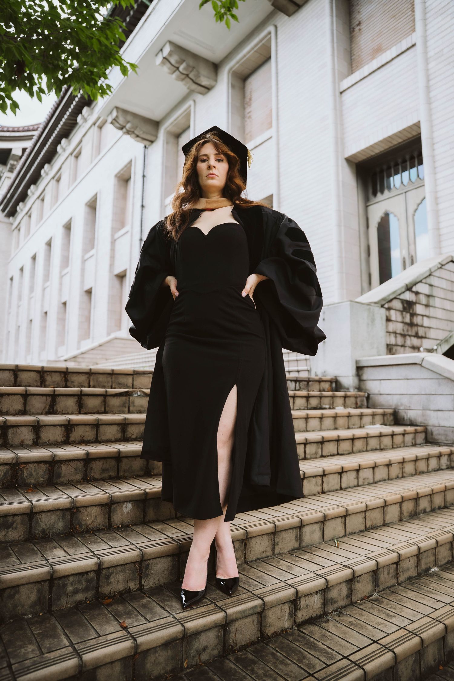 Graduate in black academic dress poses on white stone steps surrounded by greenery.