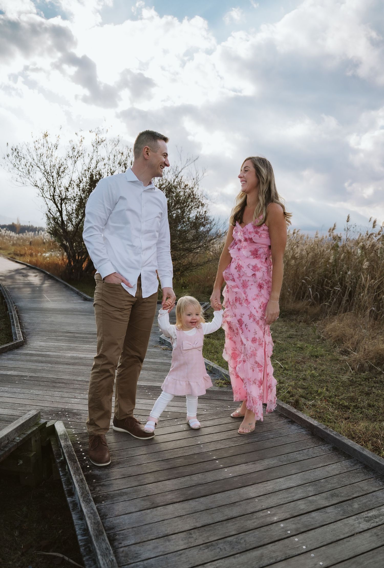 Family walking together on a wooden boardwalk through natural coastal landscape under cloudy skies.