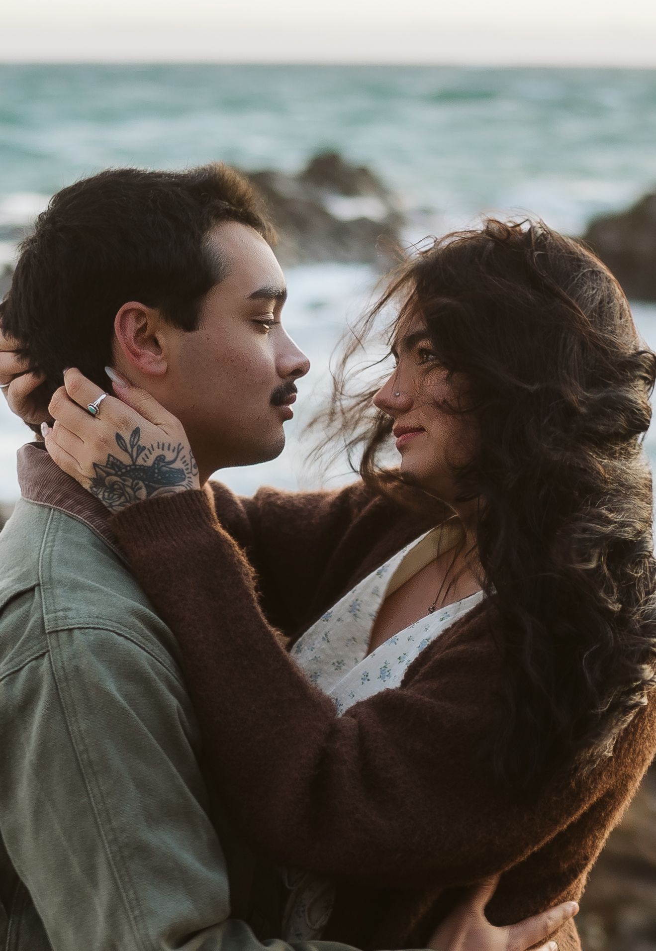 A romantic couple embraces on a windy beach at sunset as their hair blows in the coastal breeze.