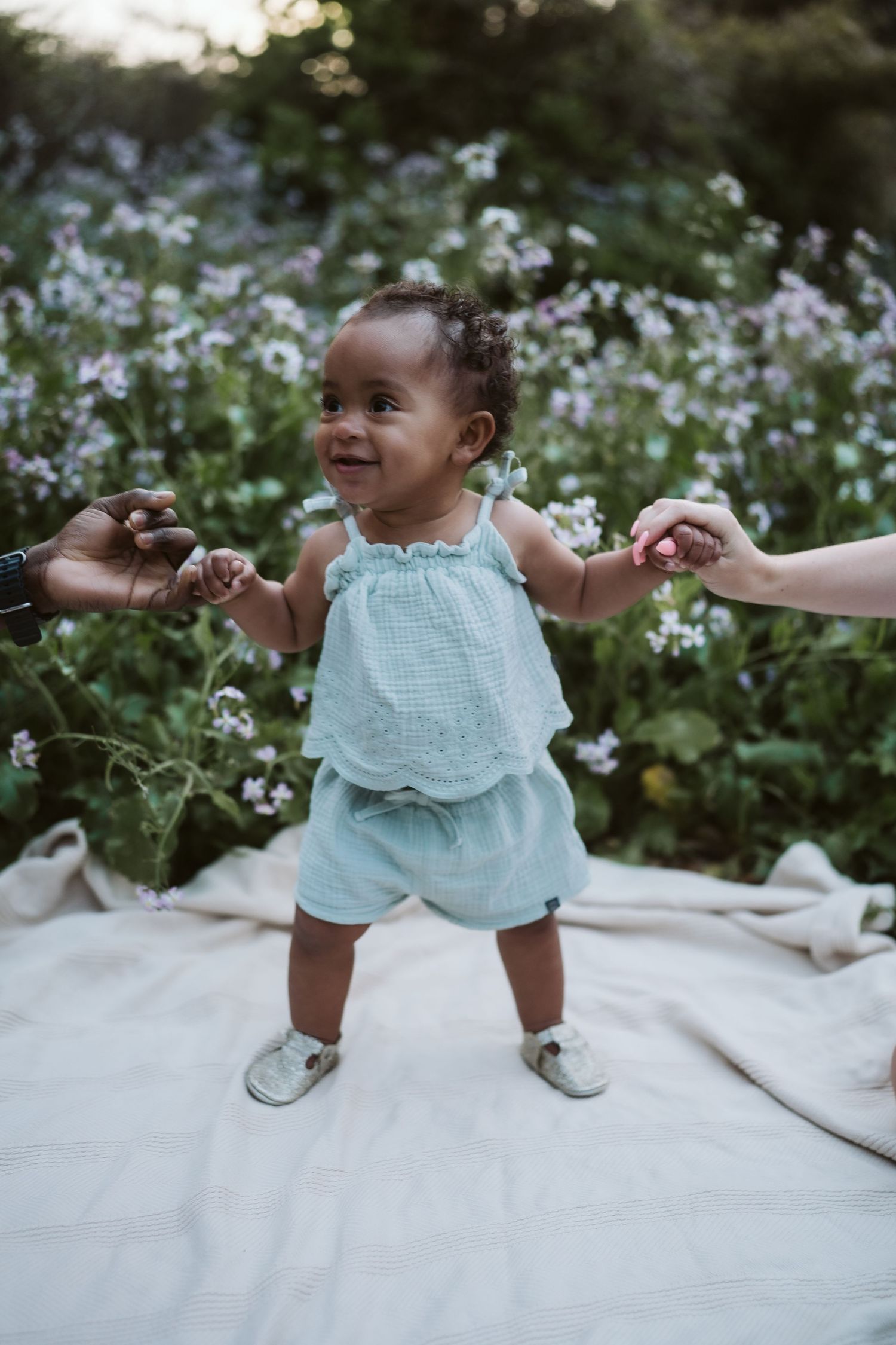 A baby in light blue clothing plays happily on a white blanket surrounded by wildflowers.
