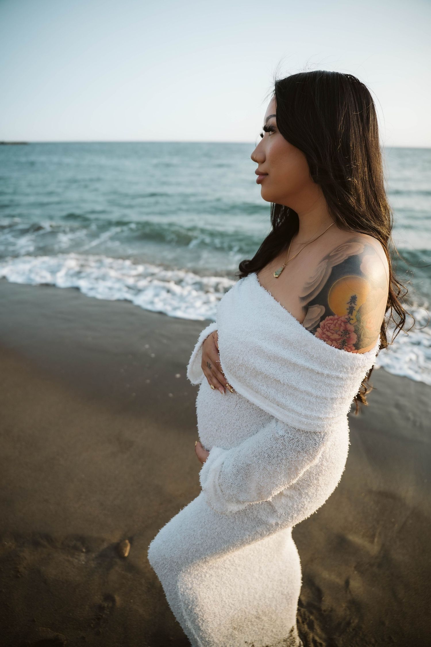 Pregnant woman in white dress poses on beach at sunset showing her baby bump.