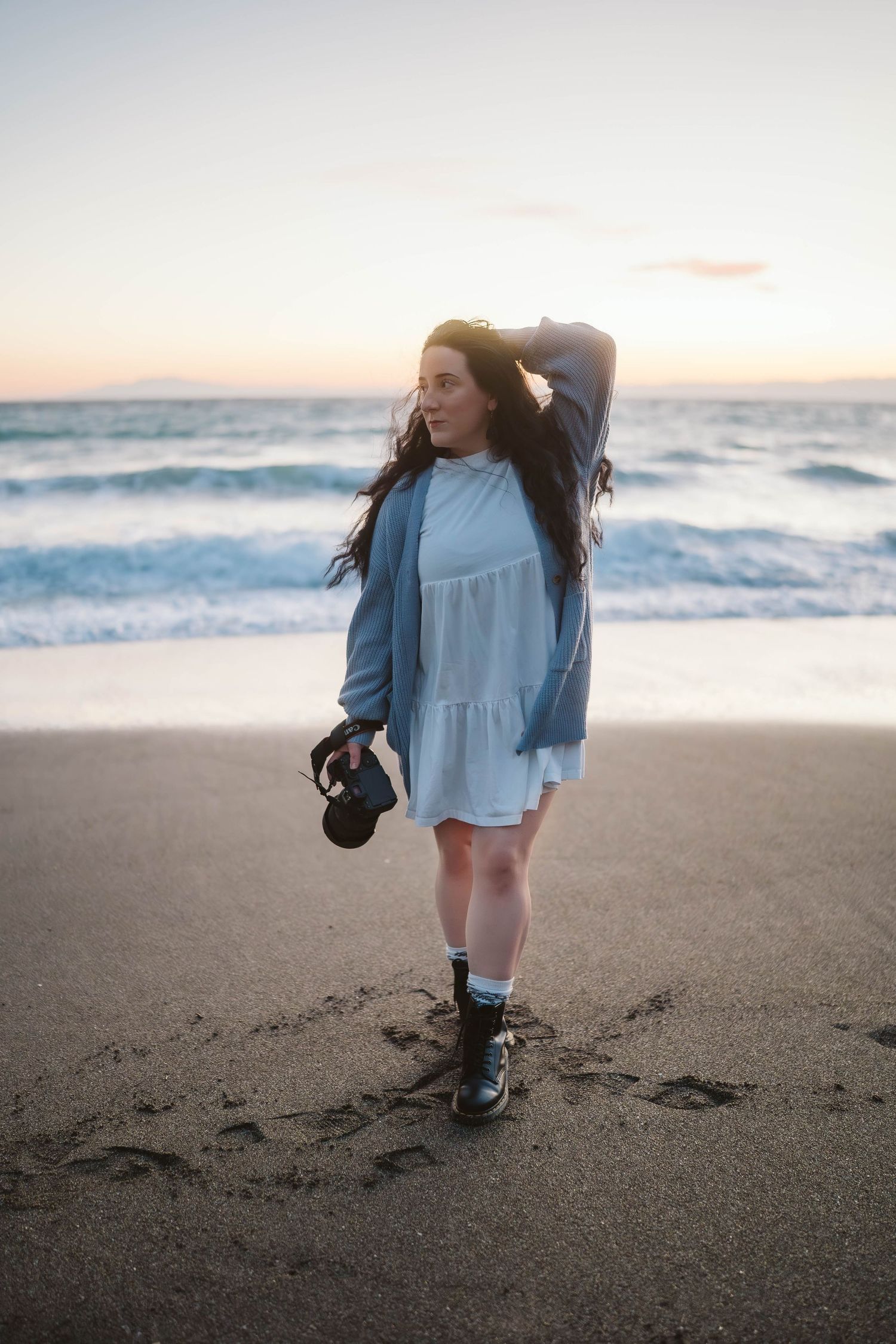 A photographer stands on a sandy beach at sunset wearing a blue dress and boots while holding a camera.