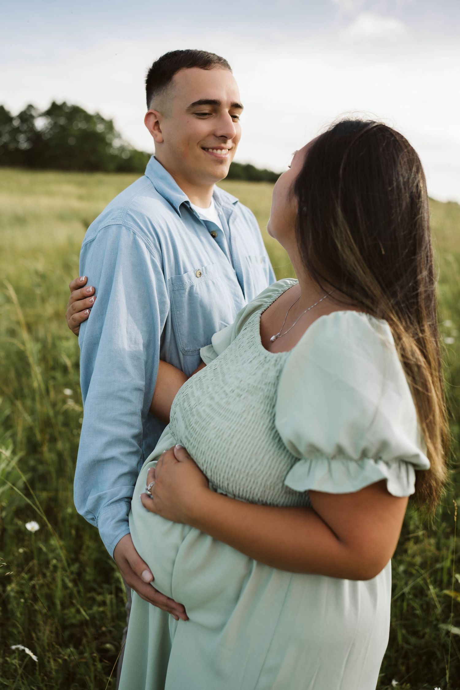 Couple embraces in a serene meadow at sunset, with her wearing a mint green dress and him in a light blue shirt.