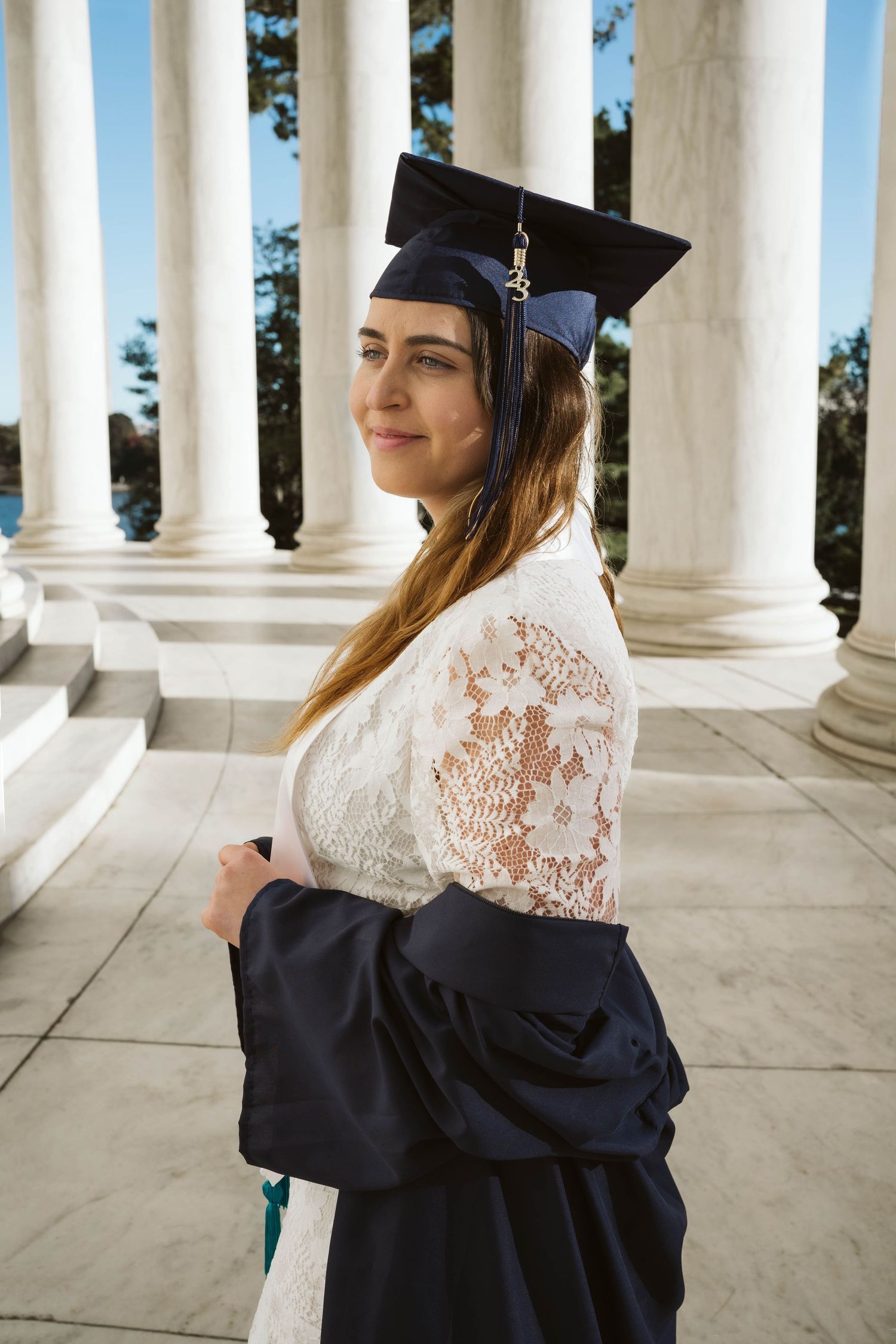 Graduate wearing black cap and gown with white lace top poses by white classical columns on sunny day.