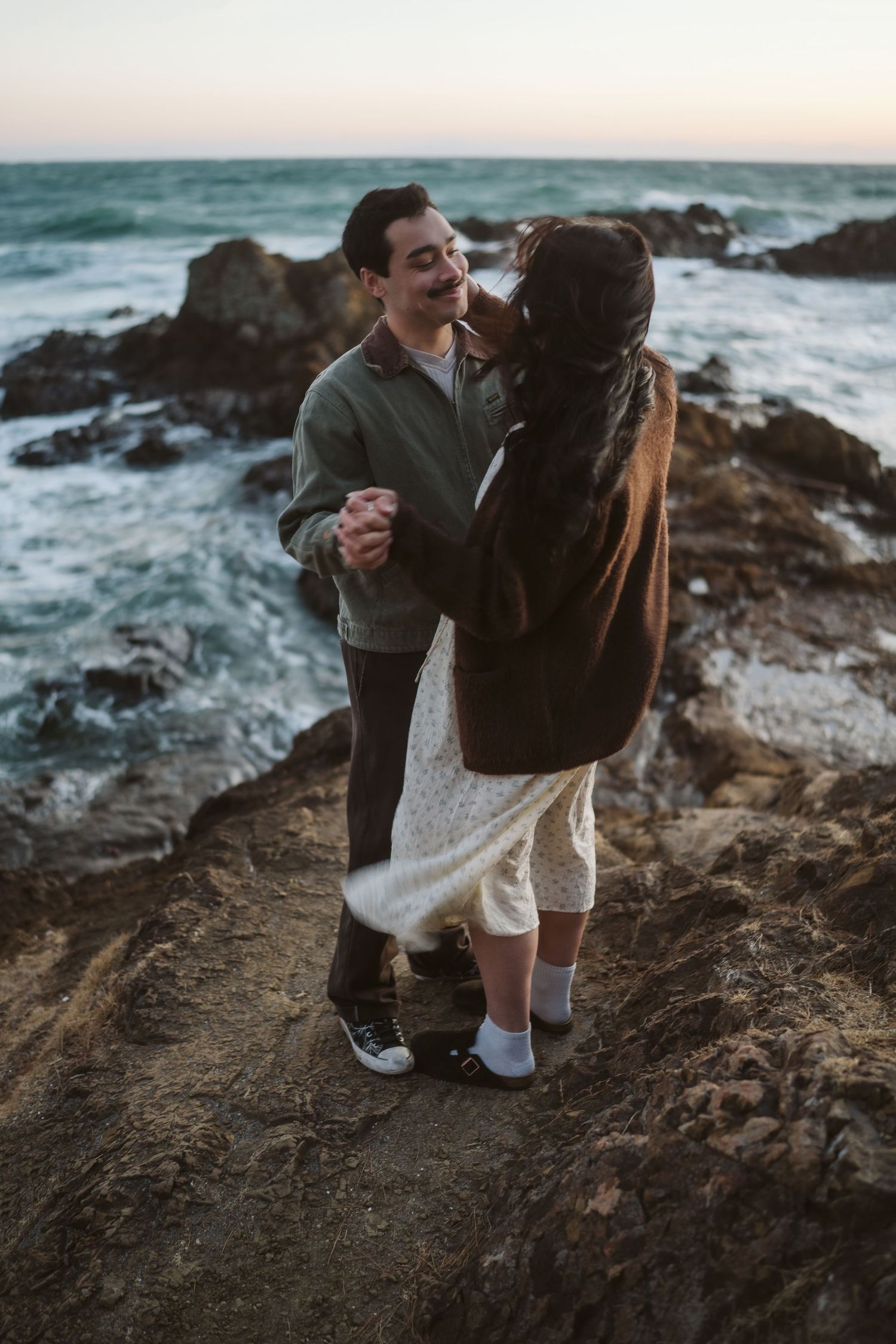 A romantic couple embraces on rocky coastal cliffs at sunset with crashing ocean waves in the background.