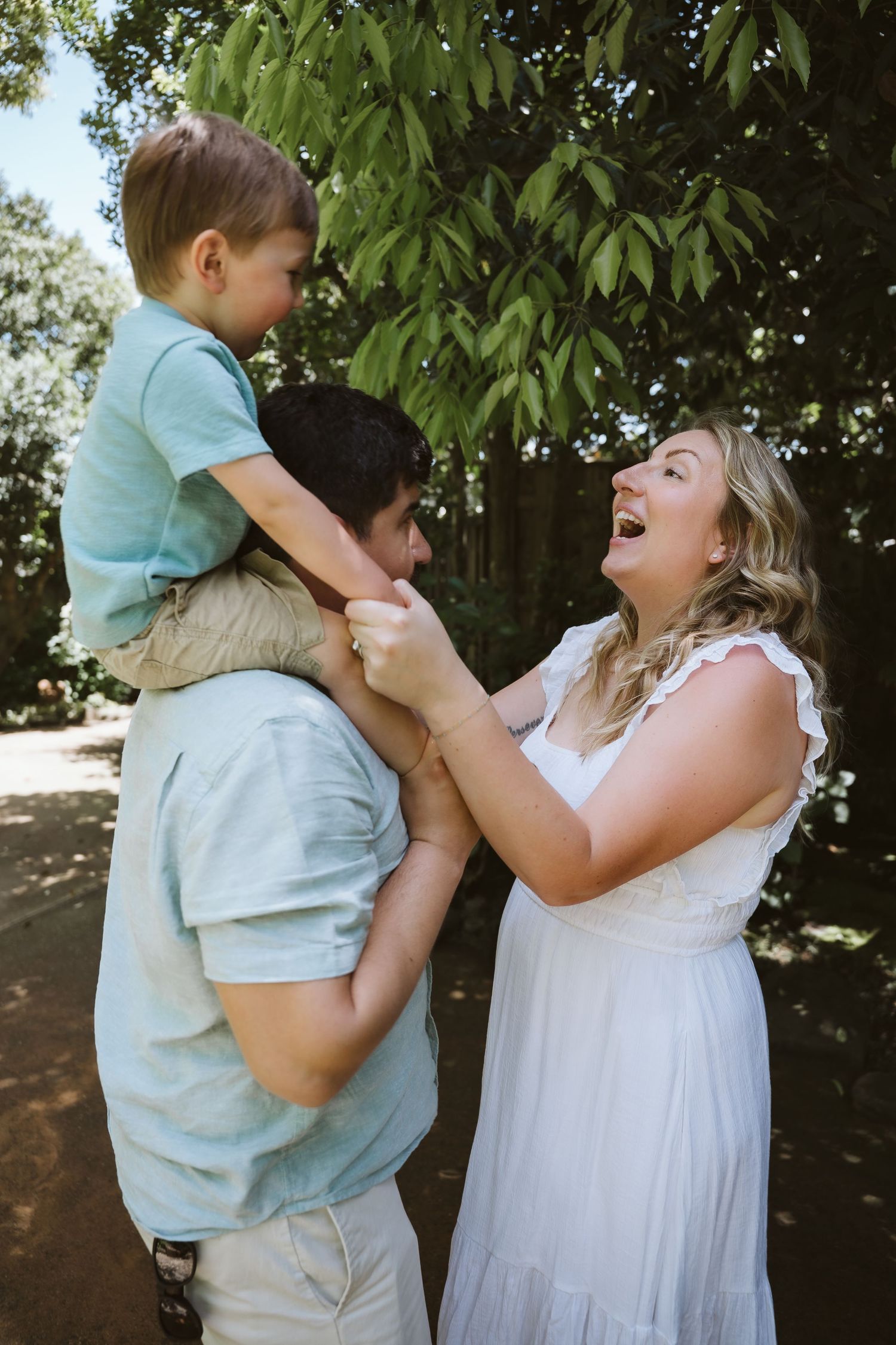 Playful family moment captured outdoors under green leafy trees on a sunny day.