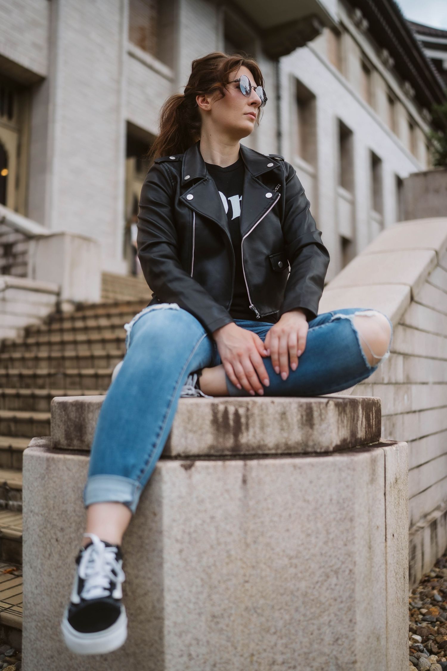 Person in leather jacket and jeans sits casually on concrete steps wearing sneakers.