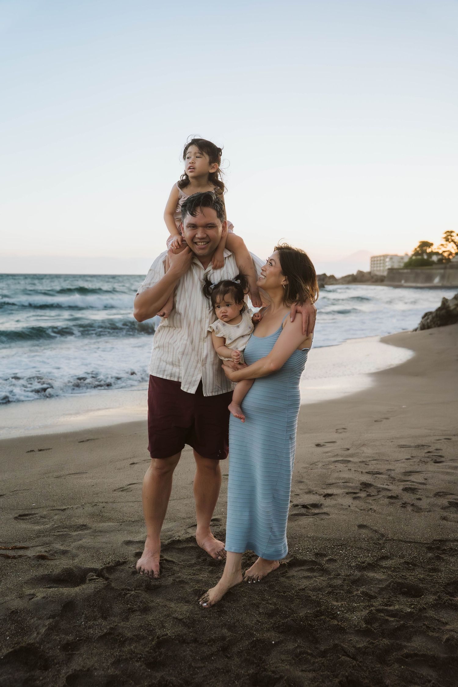 A family shares tender moments together on a black sand beach at sunset with ocean waves in the background.