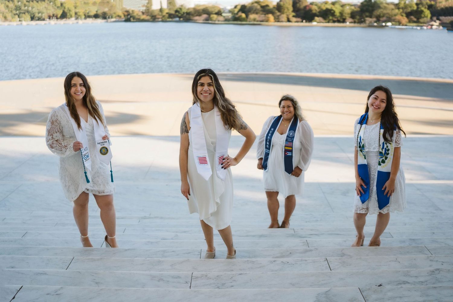 Four people in denim and white outfits pose together on a waterfront promenade near a lake on a sunny day.