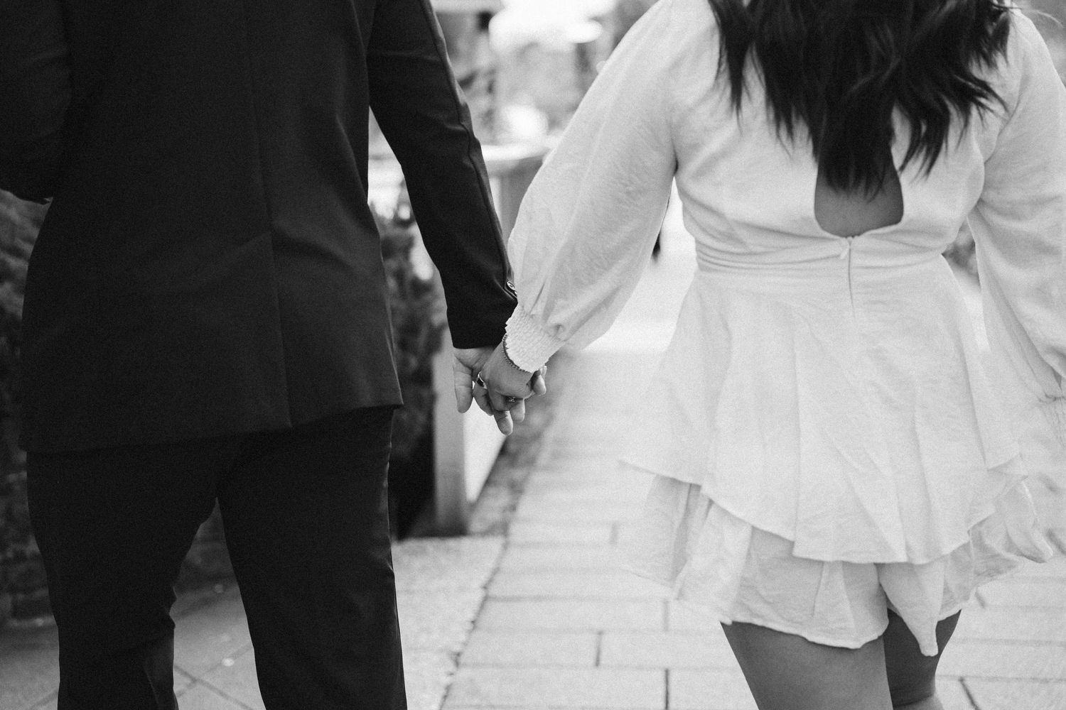 A couple holding hands walking down a stone path, with the bride wearing a short white dress and groom in black suit.