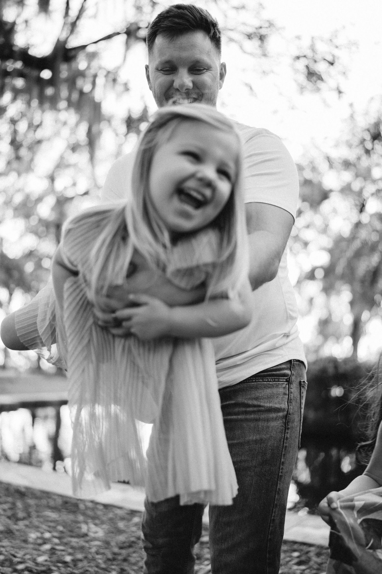 A joyful black and white candid moment shows a tender embrace between an adult and laughing child outdoors.