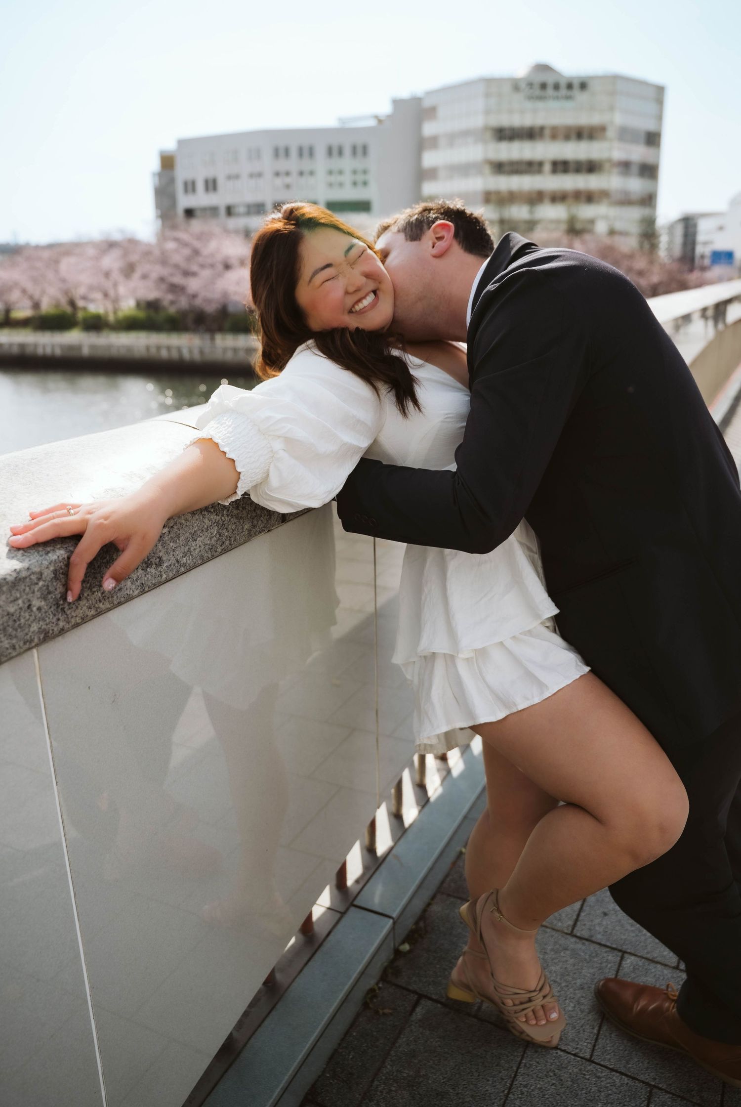 A couple shares a romantic moment on a bridge with cherry blossoms and a building in the background.