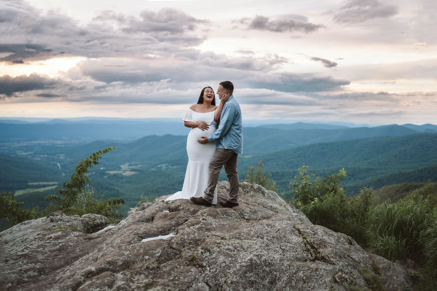A couple in wedding attire stands atop a mountain peak overlooking a vast mountain range during sunset.