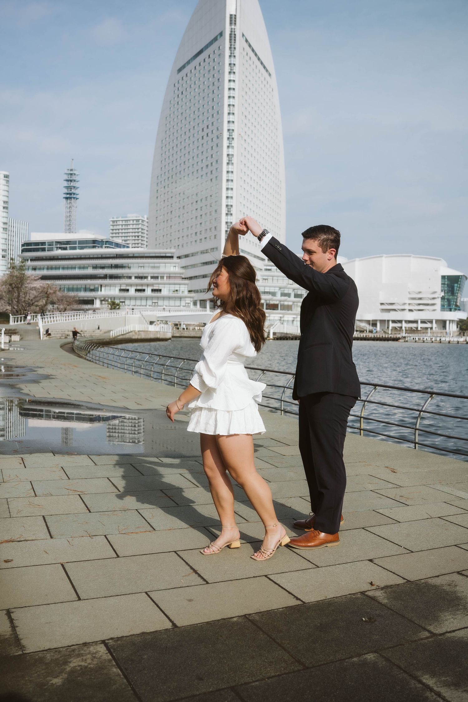 A couple dancing together on a waterfront promenade with a modern high-rise building in the background.