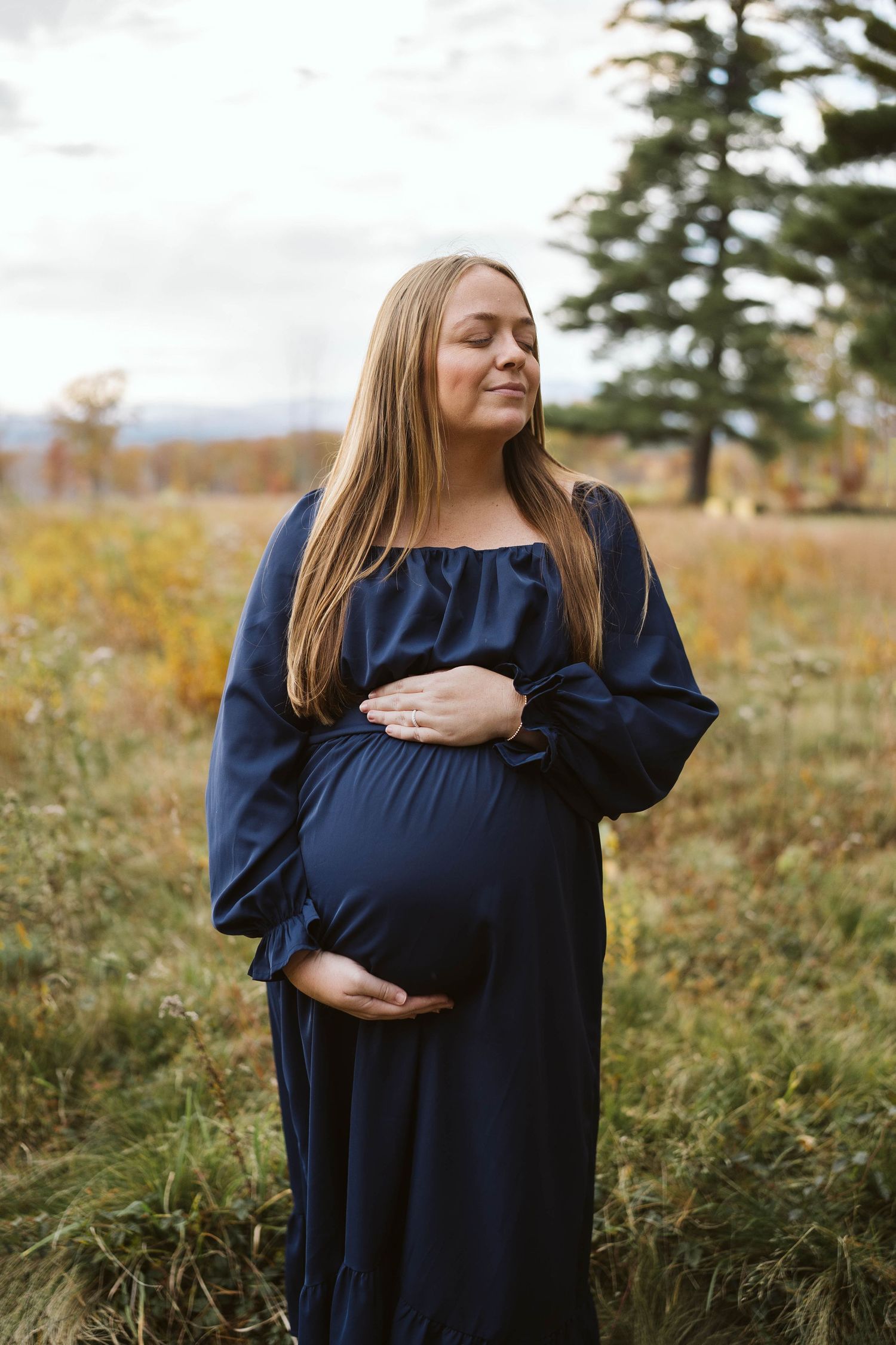 Expectant mother in navy dress poses serenely in autumn field with evergreen trees behind her.
