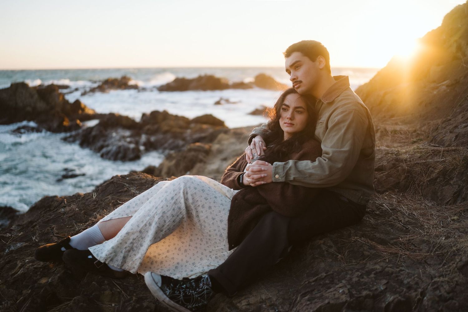 A couple embraces while sitting on coastal rocks during sunset with ocean waves in the background.